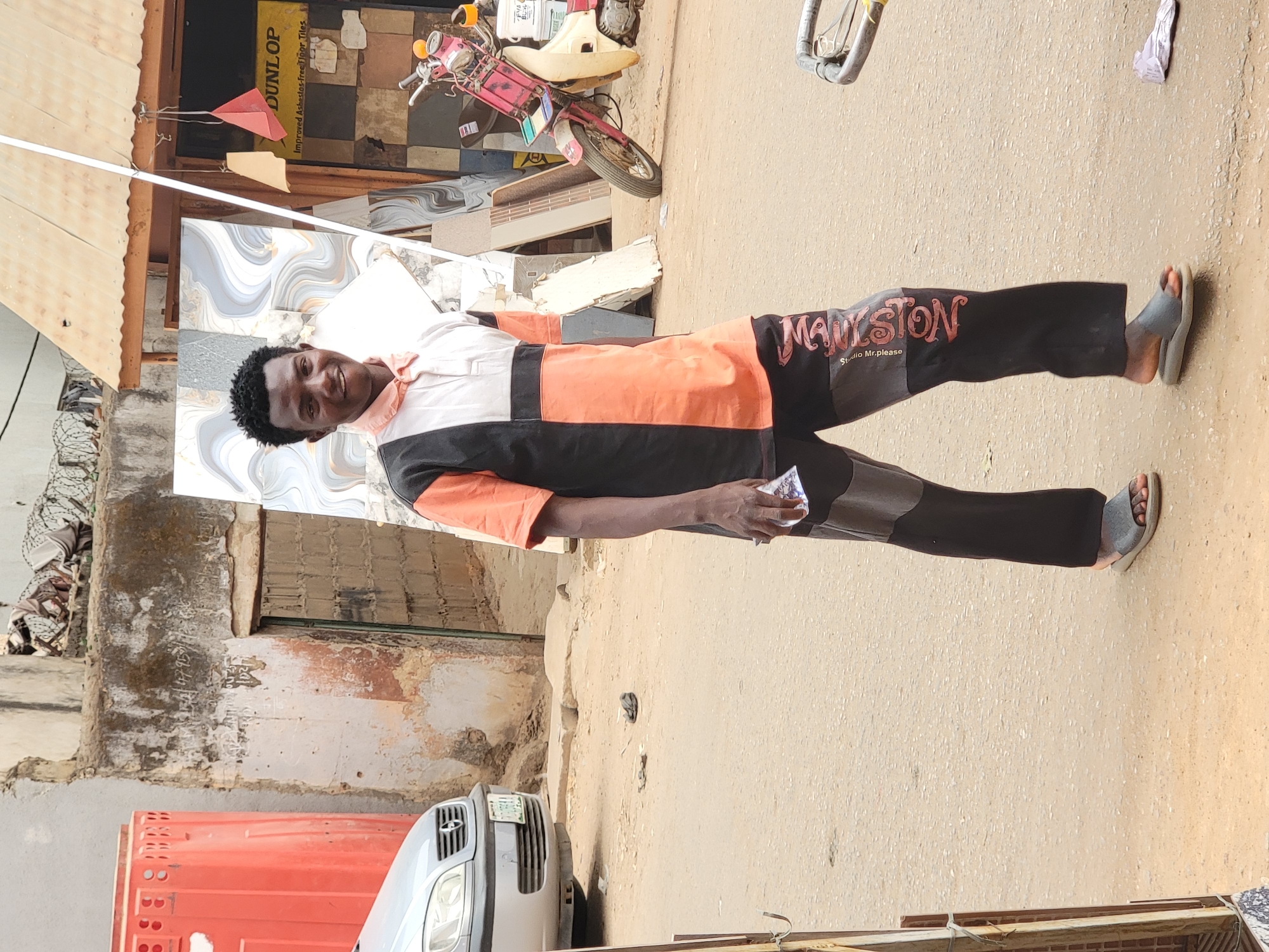 A man stands in the street, smiling at the camera. He wears a color-blocked shirt of white, orange, and black, and dark pants with "MAYLSTON Studio Mr.please" printed on the leg. His feet are clad in grey sandals. The street appears unpaved and dusty, with a car parked on the left. There is a red container, and a building with a tiled facade. A motorcycle is in the background. The location is Maiburiji, Nigeria.