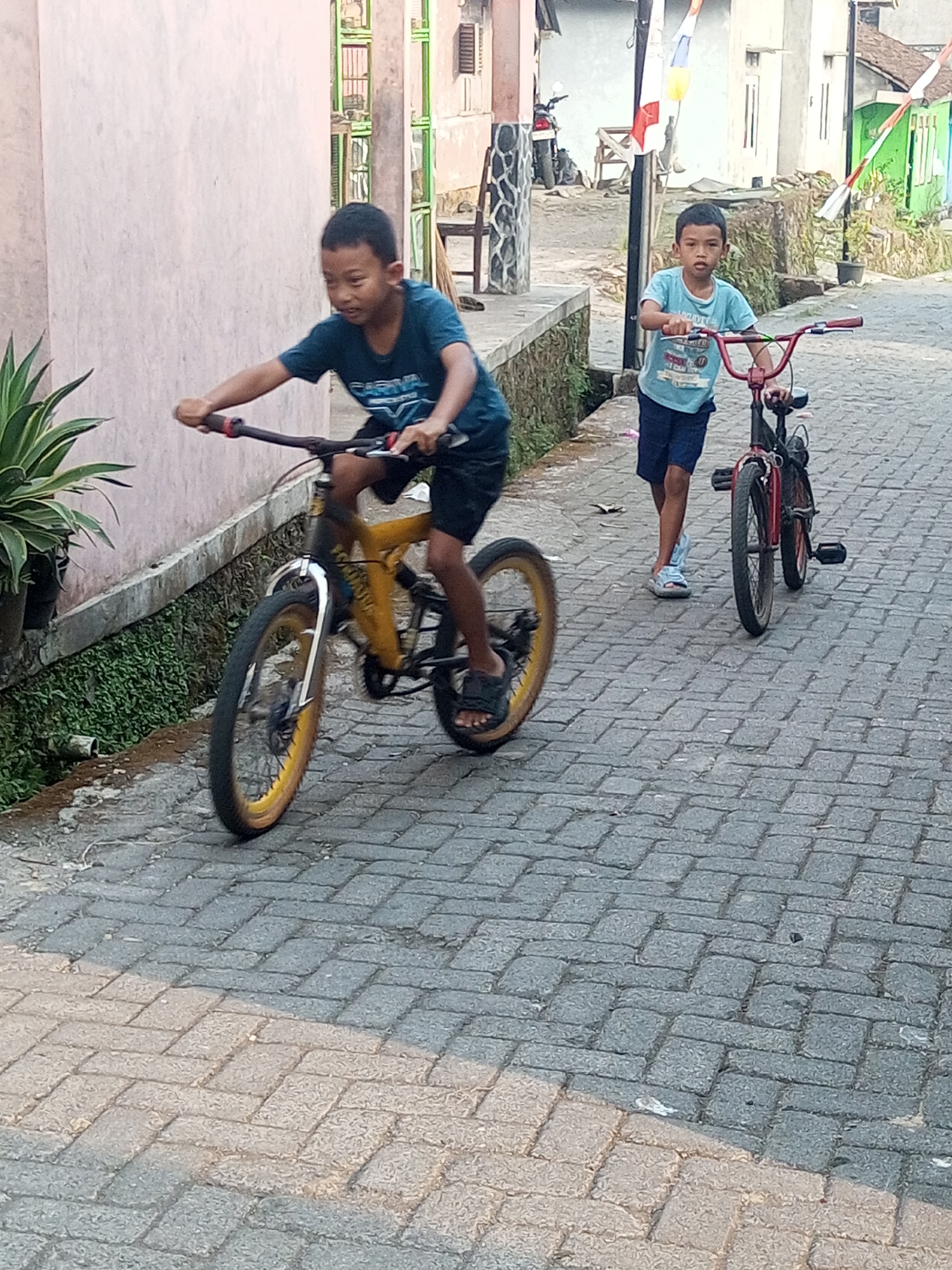 In Andongsili, Indonesia, two boys are riding bicycles on a paved street. The boy in the foreground, wearing a dark blue t-shirt and black shorts, is on a yellow and black bicycle with yellow rims. He is actively pedaling and appears to be in motion. To his right and slightly behind, another boy in a light blue t-shirt and dark blue shorts is on a red bicycle, also appearing to be riding. The street is paved with interlocking bricks, and there is a wall on the left side of the image, with some greenery and plants. In the background, buildings and a motorcycle are visible, suggesting a residential area. The scene captures a moment of childhood activity on a typical day.