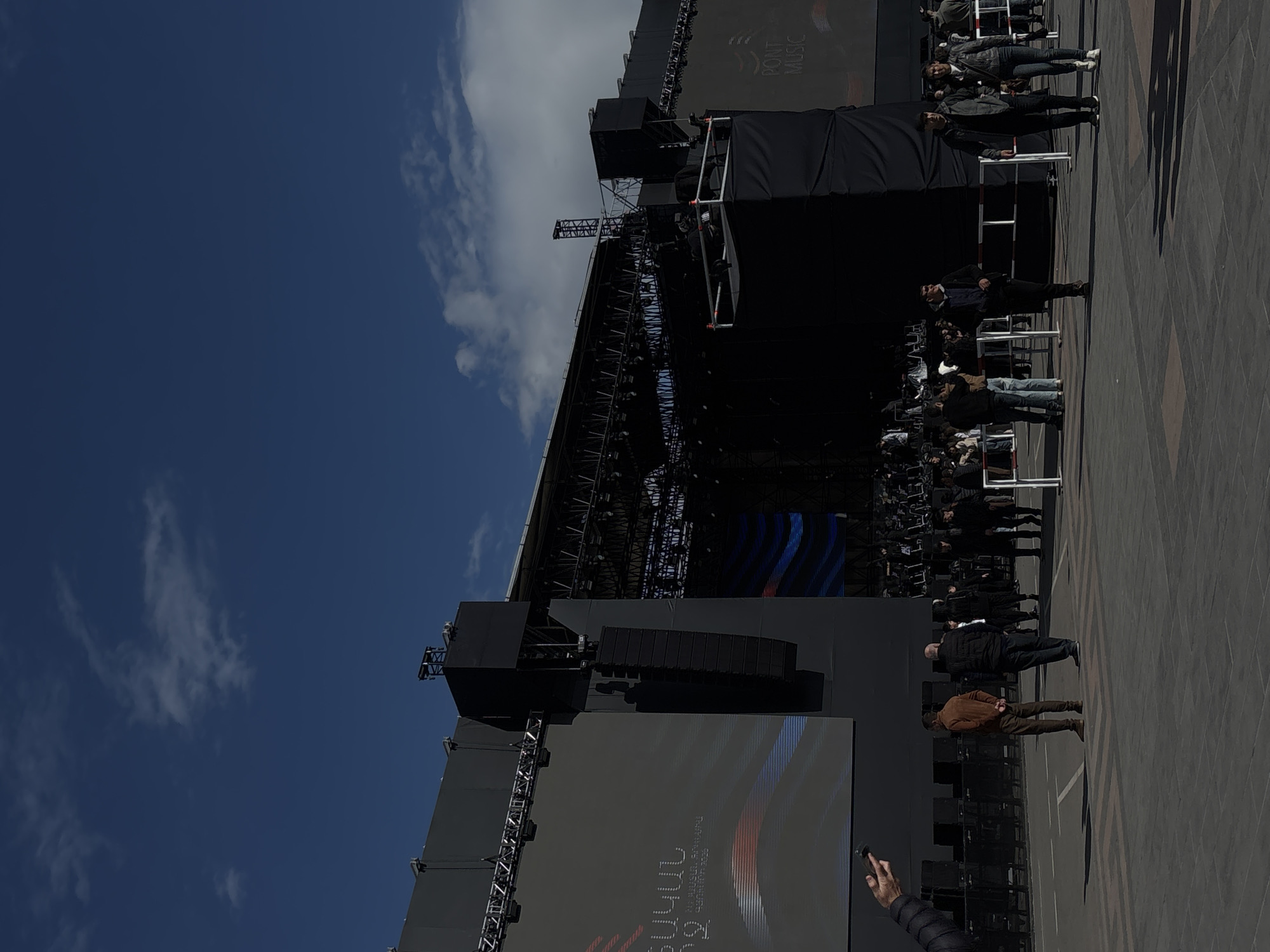 The image shows a large outdoor stage set up in what appears to be the city of Gyumri, Armenia, based on the visible script. The stage features large screens displaying visuals. A crowd of people is gathered in front of the stage, some standing on raised platforms or barriers. The background includes a blue sky with some clouds. The scene seems to be from an event or performance taking place in a public space.