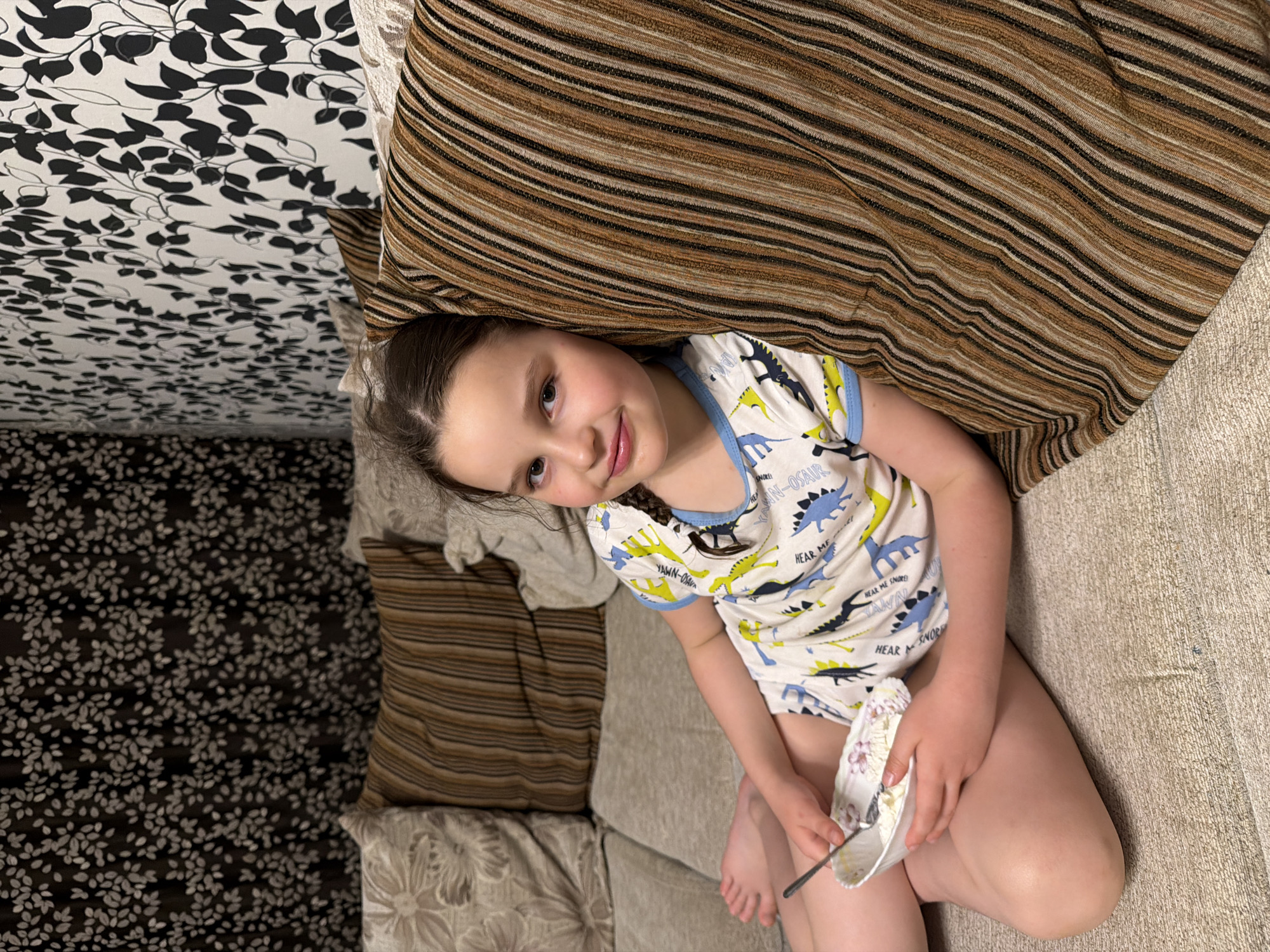 A young girl is pictured lying on a couch indoors. She is wearing pajamas patterned with dinosaurs and is holding a small bowl with a spoon in it. Her hair is braided. A striped pillow is partially covering her head. The couch has cushions with different patterns, including a floral one. The wall behind the couch is decorated with a leaf-patterned wallpaper. The location in Zaprudnya, Russia, is not discernible from the image.