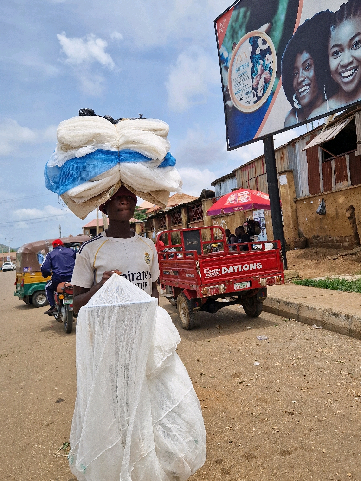 In a dusty outdoor marketplace in Unknown, Unknown, a young person walks forward, carrying a large load of white and blue netting on their head. They are wearing a white t-shirt with a Real Madrid logo and "Emirates" written on it. In their hands, they hold a white mosquito net. In the background, a red truck is parked on the side of the road, with people sitting in its bed. A billboard advertising "ZYN BUTTER" with images of Black women is visible above a row of corrugated metal structures. An umbrella shades a small stall. A three-wheeled vehicle is also visible on the road, with a person in a red hat riding on the back. The ground is unpaved and scattered with debris. The sky is partly cloudy.