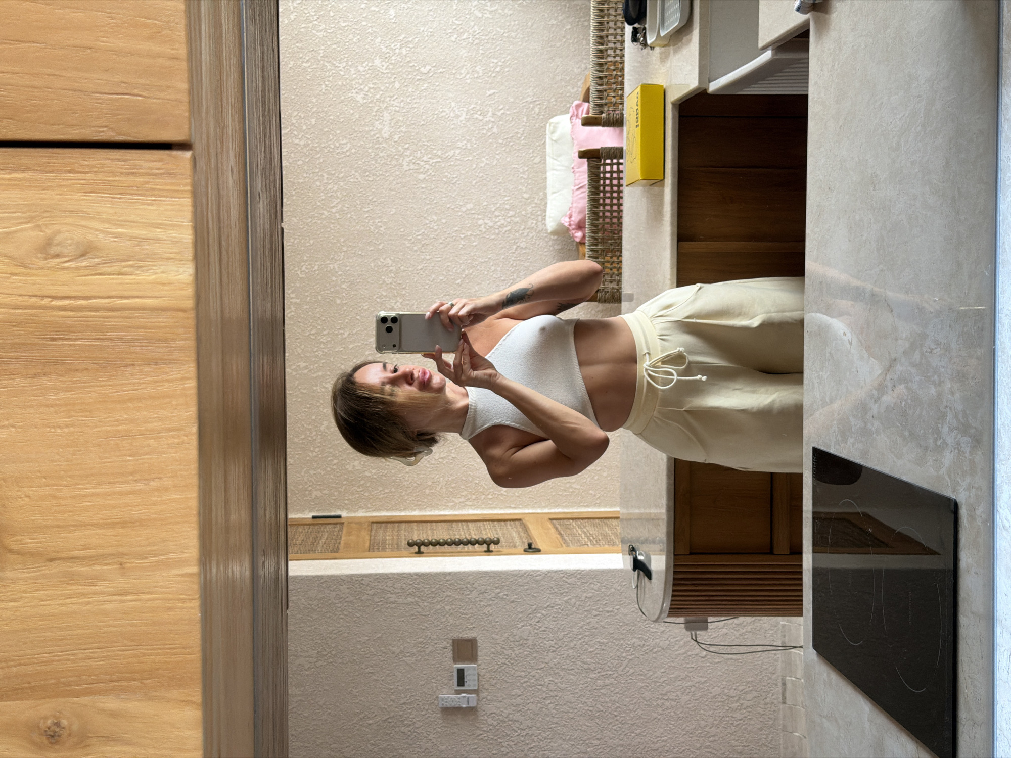 A woman stands in a kitchen and takes a mirror selfie. She is wearing a white crop top and light-colored pants. The kitchen features wooden cabinets above and a countertop with a built-in stovetop. In the background, there is a sink, a yellow box, and a woven chair with pillows. The location is the city of Mengwi, Indonesia.