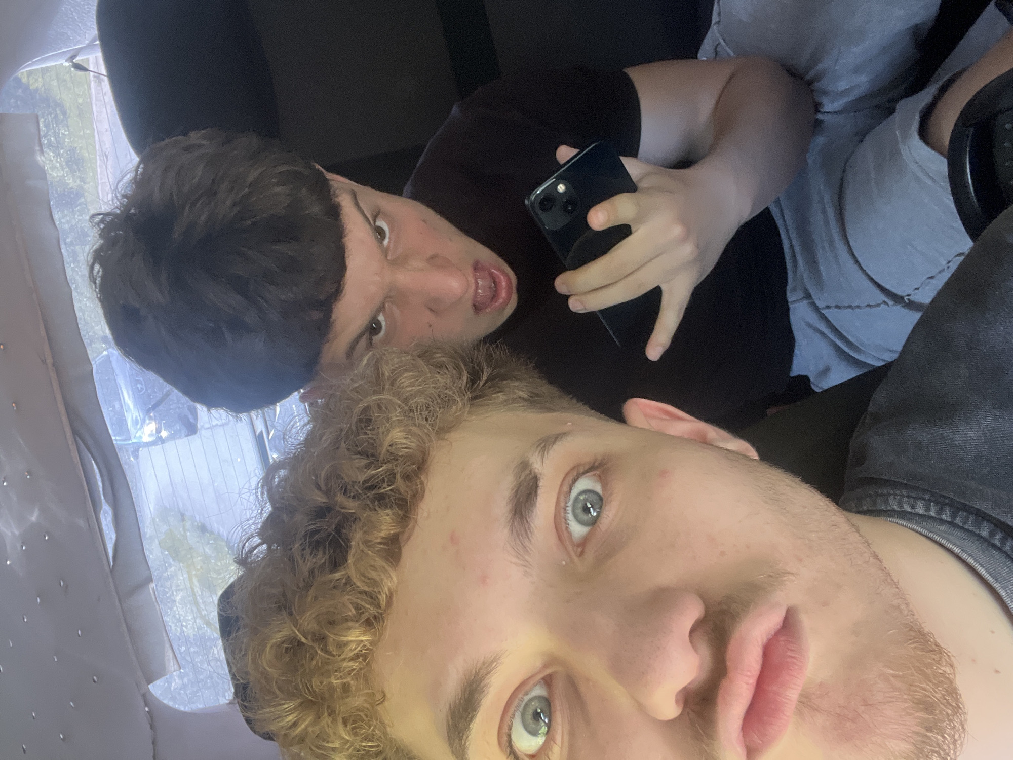 The image shows two young men inside a vehicle, possibly a car. The man in the foreground, on the left, has curly blond hair and light-colored eyes. He is looking directly at the camera with his mouth slightly open. The man behind him, on the right, has short, dark hair and is making an angry or surprised facial expression, with his mouth open and teeth visible. He is holding a dark-colored smartphone in his right hand. Both individuals are wearing casual clothing. The interior of the vehicle is visible, with a headliner in the background that has small reflective elements. The rear window of the vehicle is also visible, showing a blurred outdoor scene with trees and what appears to be a building or structure. The image is taken from a low angle. The specific location in Vlahovo, Bulgaria, cannot be determined from the visual information provided in the image.