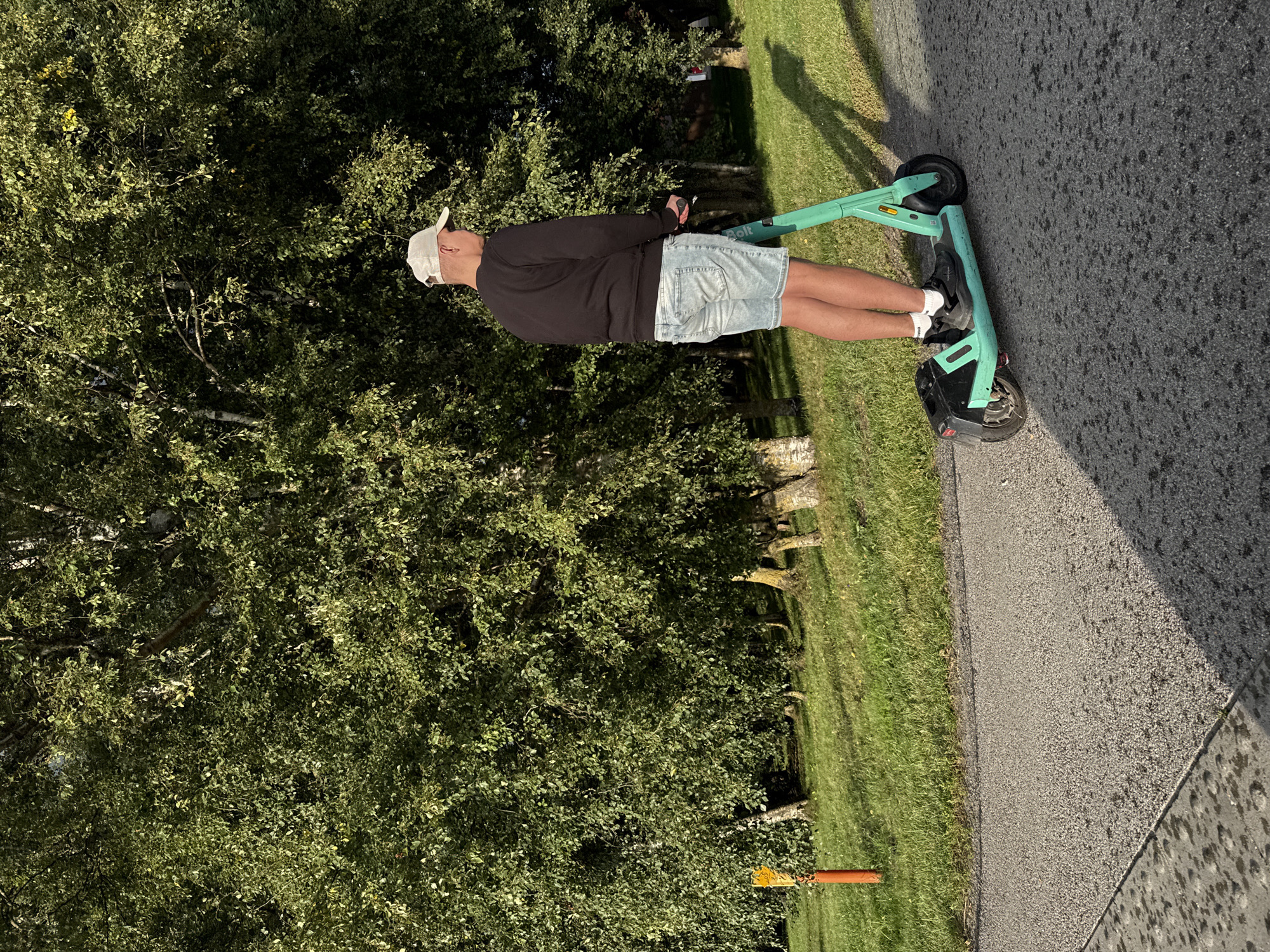 A man is riding a teal electric scooter on a paved pathway. He is wearing a black long-sleeved shirt, light blue denim shorts, a white baseball cap, white socks, and black sneakers. The scooter has black accents and a black rear fender. The pathway is surrounded by a grassy area with several trees. A bright orange marker with yellow markings is visible in the grass to the left. The scene appears to be a park or a similar outdoor recreational area. The lighting suggests it is late afternoon or early evening, with long shadows cast by the trees. There is no visible indication of the specific location within Tallinn, Estonia.
