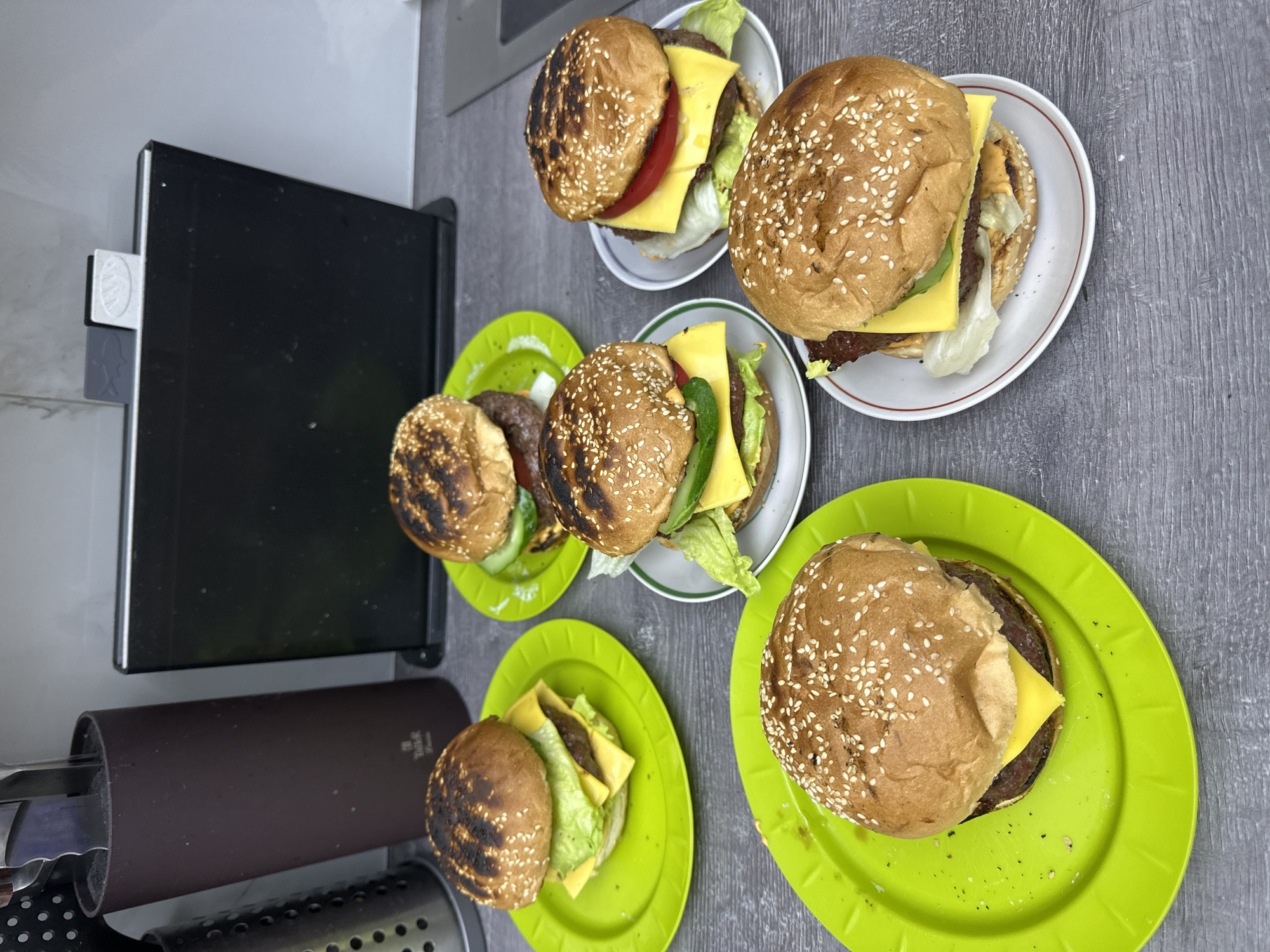 The image displays six homemade hamburgers arranged on a gray, wood-textured surface, likely a kitchen countertop or table, in слобода Старая, Russia. Three of the hamburgers are placed on bright lime green plastic plates, while the remaining three rest on small white ceramic plates with colored rims (red or green). Each burger consists of a sesame seed bun, a patty, a slice of yellow cheese, and fresh lettuce. Several also include slices of red tomato and green cucumber. The buns exhibit varying degrees of toasting, with some showing distinct char marks, suggesting they were recently grilled.
On the left side of the frame, a black electric grill or griddle is partially visible, likely the cooking surface for the burgers. Attached to its side is a small white and gray holder, featuring a stylized fish or dolphin symbol on its lower part. Below the grill, a dark brown cylindrical knife block, with a visible "TAIER" logo, is partially seen, along with a black, perforated utensil holder. The upper left background shows a plain light gray wall or cabinet. The scene depicts a casual home meal setting, likely during daytime, centered around the freshly prepared hamburgers.