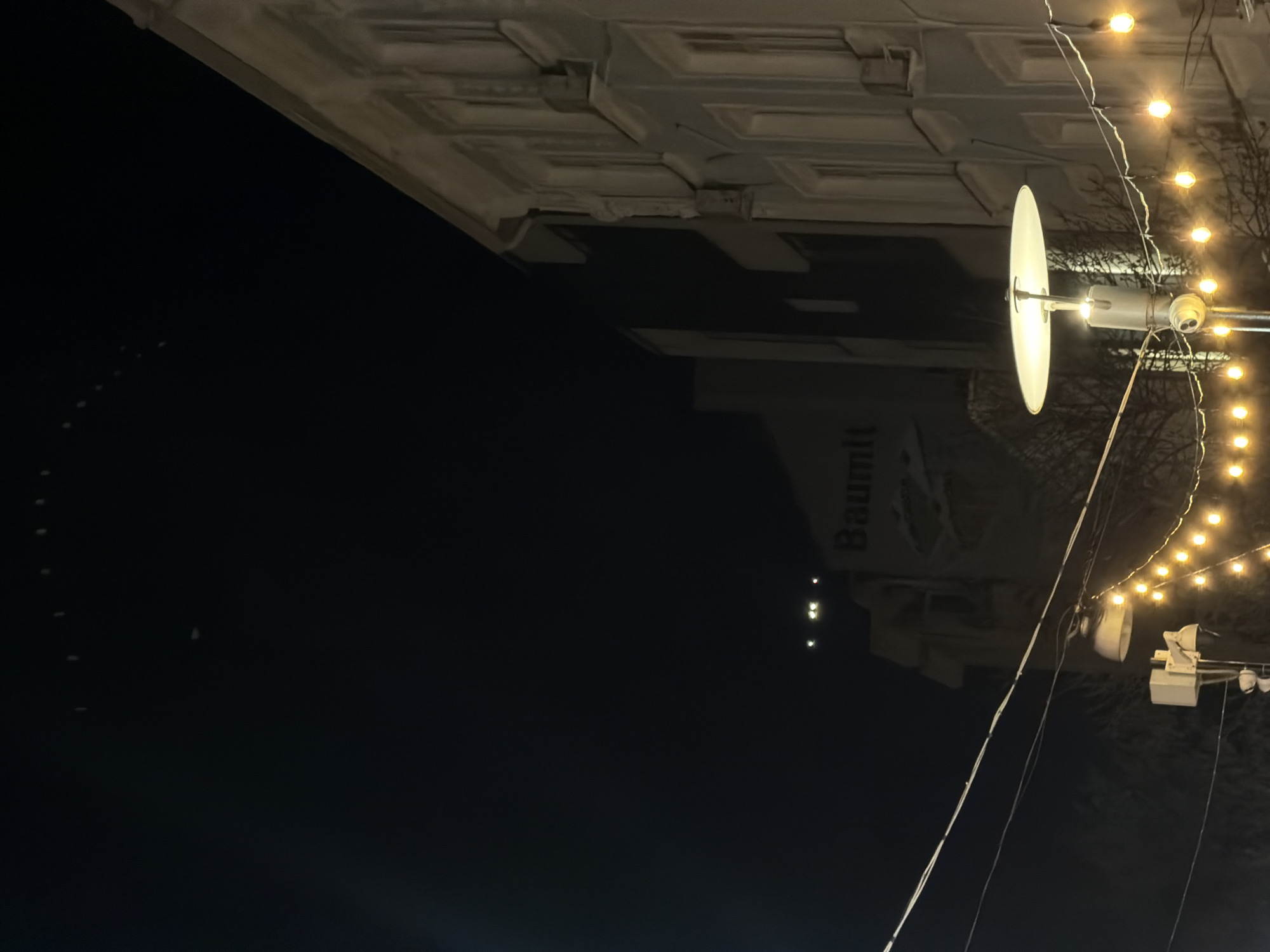 The image shows a night scene with a building at the top and dark sky below. A street lamp is visible in the upper right, strung with lights. A large building or structure is present in the upper portion of the image with the word "Baumit" printed vertically. The location is Unknown, Unknown.