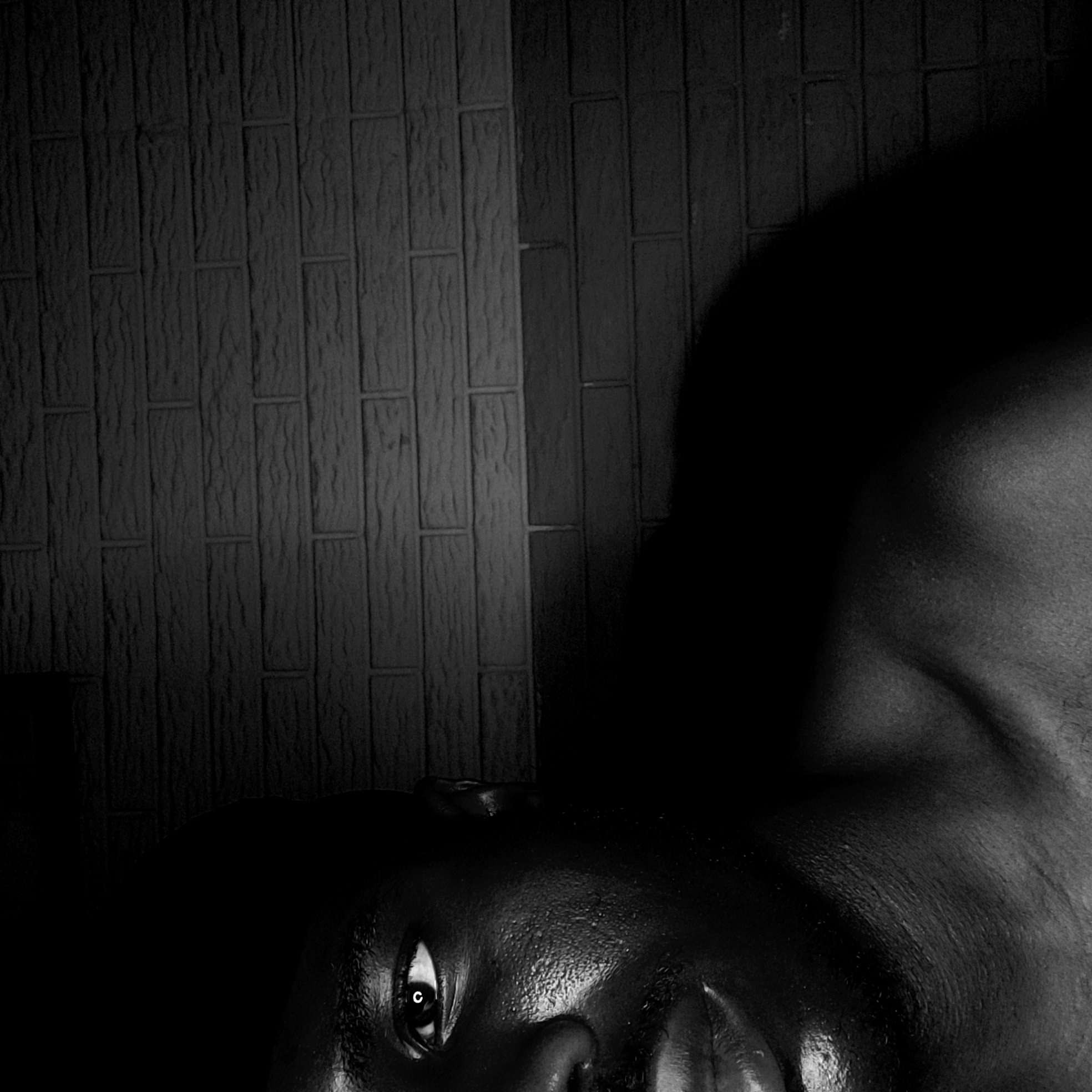 A black and white photograph shows a person lying down next to a brick wall. The person is lying on their side with their head closer to the bottom of the frame and their body extending toward the top right corner. Only one eye is visible. The location is not specified as Narayi, Nigeria, in the image.