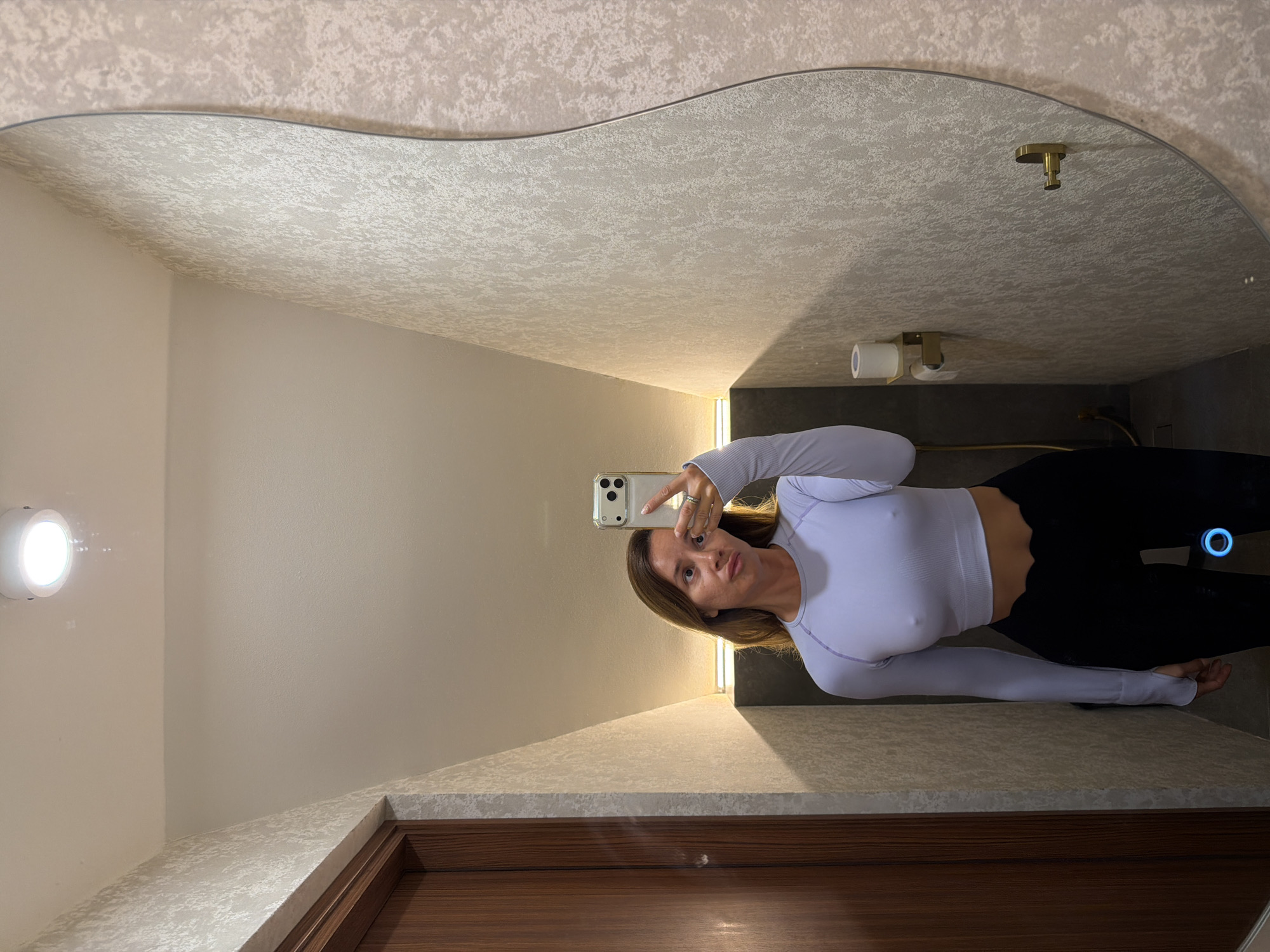 A woman in workout attire takes a mirror selfie in a restroom. She is wearing a light purple long-sleeved crop top and black leggings. The restroom has textured wallpaper, a wooden door, and a large, wavy mirror. There is a toilet paper holder with a roll of toilet paper mounted on the wall. The lighting appears to be from an overhead fixture and a strip light.