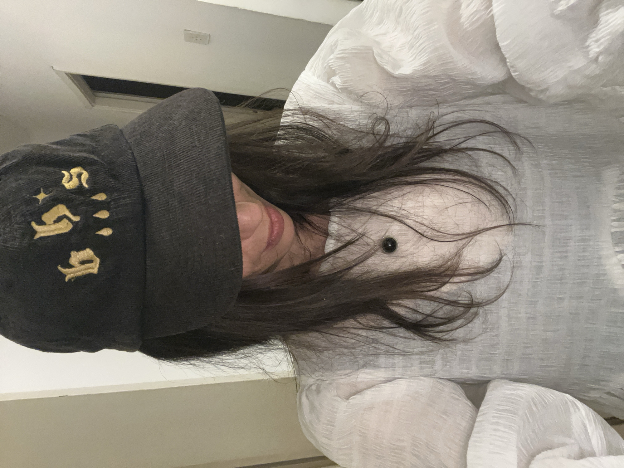 A woman wearing a dark baseball cap with gold lettering and a white textured top is looking down. A dark pendant necklace is visible on her chest. Her dark hair is long and falls around her shoulders. The background is blurry and appears to be an indoor hallway with a light-colored wall and a doorway. There is no discernible location in Sofia, Bulgaria.