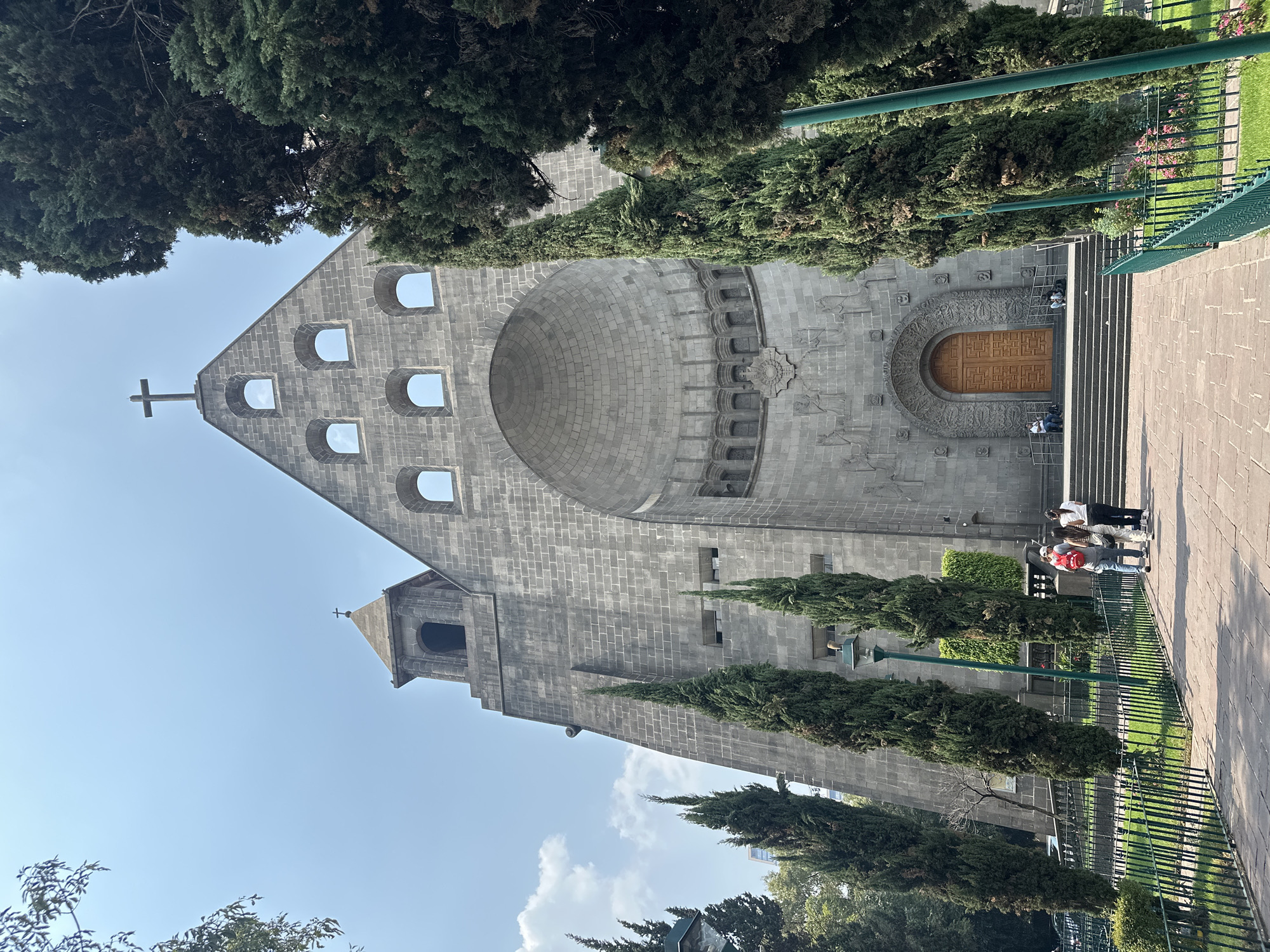 The image shows the Templo Expiatorio a Cristo Rey, also known as the Antigua Basílica de Guadalupe, in Mexico City, Mexico. The building is constructed of gray stone with a distinctive facade featuring a large, semicircular dome-like structure above the main entrance. Below the dome are a series of carvings. Above the dome, the facade continues in a triangular shape with arched windows and a cross at the peak. To the left, a bell tower rises. A flight of stairs leads up to the wooden door. A few people stand near the building's entrance. The building is surrounded by greenery, including tall, slender trees and a fenced-in park area. The sky is light blue with some clouds.