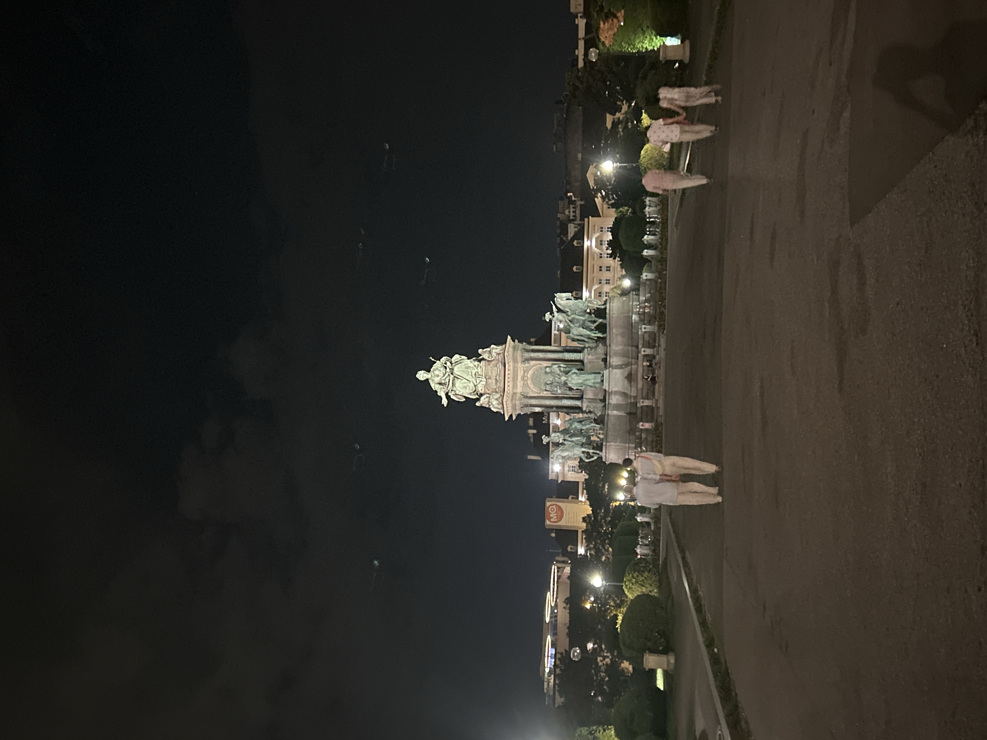 The image depicts a nighttime scene at the Maria-Theresien-Platz in Vienna, Austria. A prominent monument, the Maria Theresa Monument, stands at the center, featuring a large equestrian statue of Empress Maria Theresa surrounded by allegorical figures. The monument is illuminated, casting a glow on the surrounding area. In the foreground, a paved pathway leads towards the monument. Several individuals are visible walking away from the camera on this path. Two individuals are closer to the camera, appearing to be a couple, dressed in light-colored clothing and walking side-by-side. Further ahead, another group of three individuals, also in light-colored attire, are walking together. The background shows buildings and trees, with streetlights providing ambient illumination. The sky is dark, with some faint clouds visible, and a few distant lights suggest the presence of other structures or activities in the city.