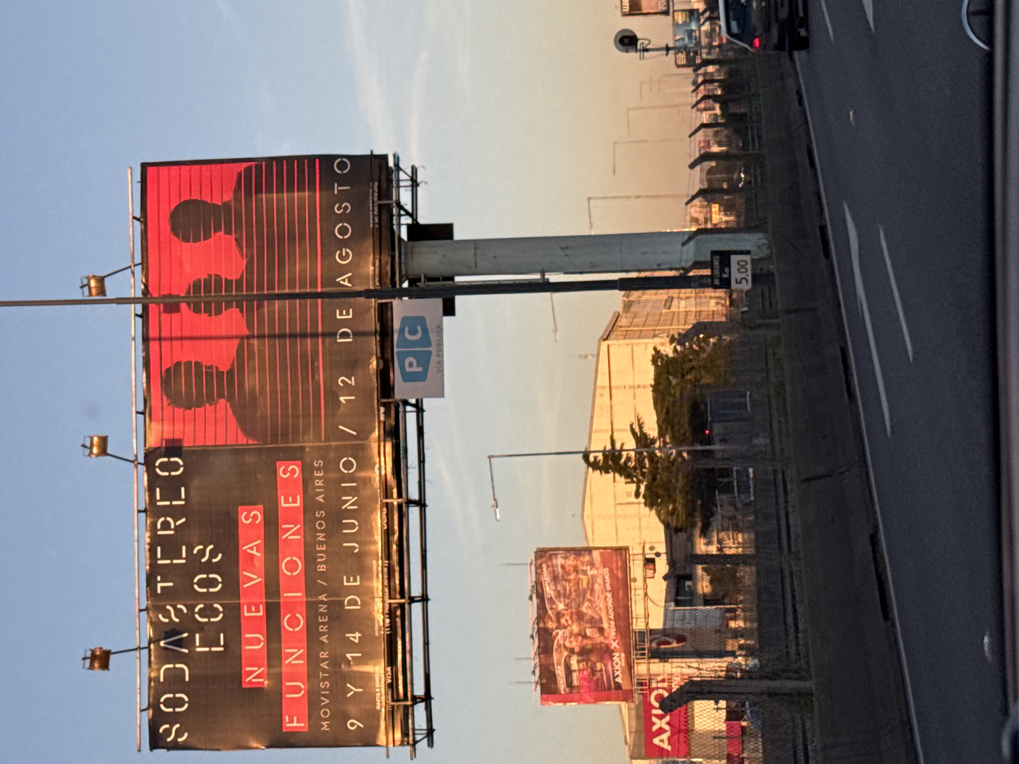 The image depicts a highway scene, with a large billboard in the foreground advertising "Soda Stereo Ecos, Nuevas Funciones" at Movistar Arena in Buenos Aires, Argentina. The billboard shows concert dates of June 9th and 14th, and August 12th. The billboard also features images of silhouettes. Further down the road, another billboard advertises "Axion". The sky is visible above the billboards. A roadside barrier runs along the edge of the highway. Several other vehicles are on the road, one of which is at the far right of the shot. There are buildings beyond the highway, as well as street lights.