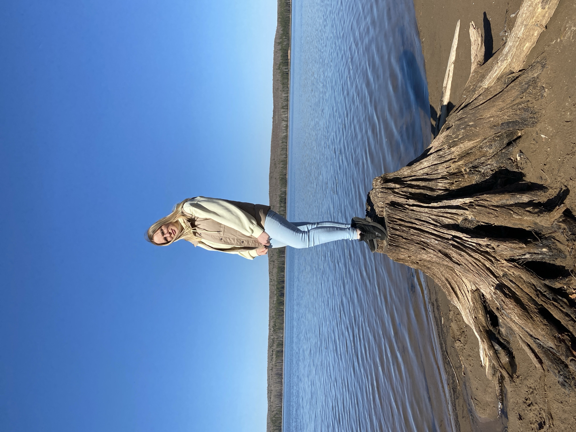 A young woman with blonde hair, wearing a beige jacket, light blue jeans, and black shoes, stands on a large, weathered tree trunk next to a body of water. The body of water appears to be a lake or river, with a visible treeline on the opposite shore. The sky is a clear, deep blue. The scene is outdoors, with sunlight present. The location is near the city of Chaikovsky, Russia.