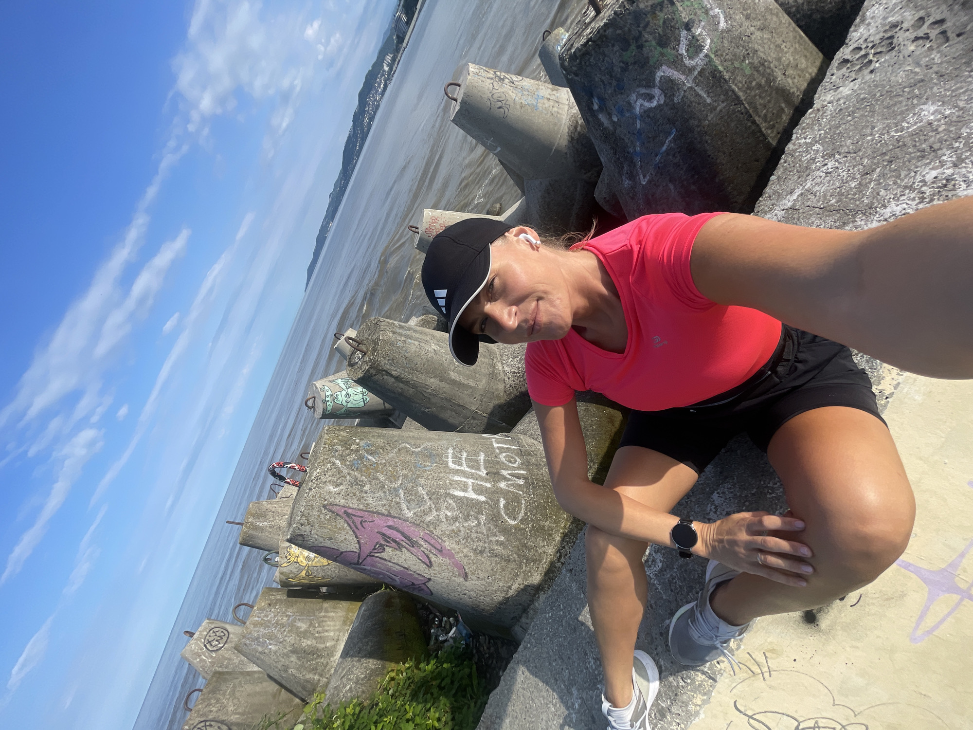 A selfie was taken on a sunny day in Sochi, Russia. The person is sitting on a concrete structure and looking at the camera. The person is wearing a pink shirt, black shorts, a black hat, white headphones, and gray shoes. The body of water and vegetation is visible in the background.