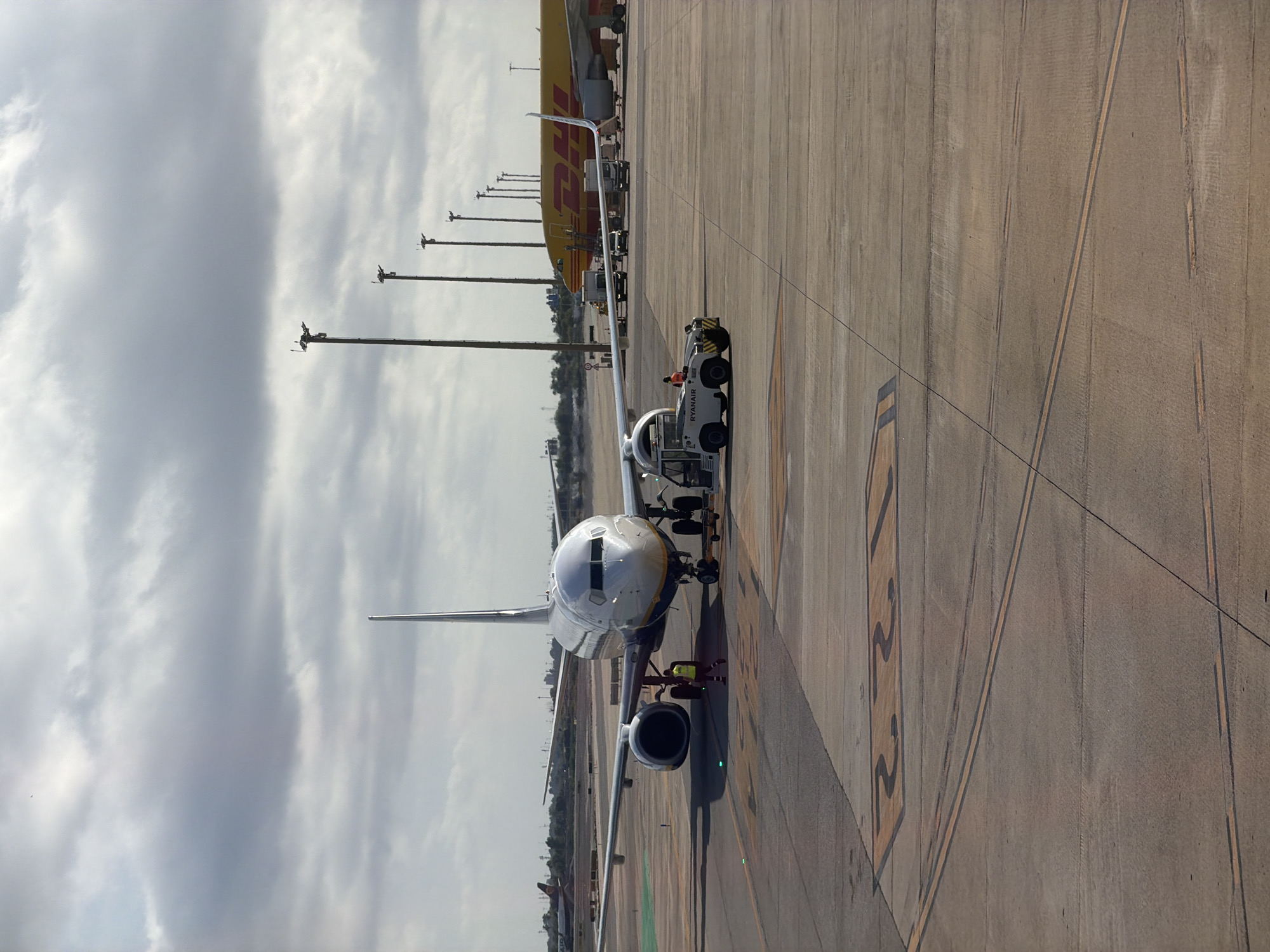 The image shows an airport tarmac with several aircraft and service vehicles. In the foreground, a Ryanair airplane is parked on the tarmac, with a ground crew member in a high-visibility vest standing near its front landing gear. A baggage tug, also labeled "Ryanair," is positioned in front of the plane, likely preparing to move it. Further in the background, a large yellow DHL cargo plane is visible, along with other vehicles and what appears to be airport lighting poles. The scene is set against a cloudy sky. The presence of Ryanair aircraft and the general airport environment suggest this could be Barcelona-El Prat Airport (BCN), as Ryanair is a major operator there.