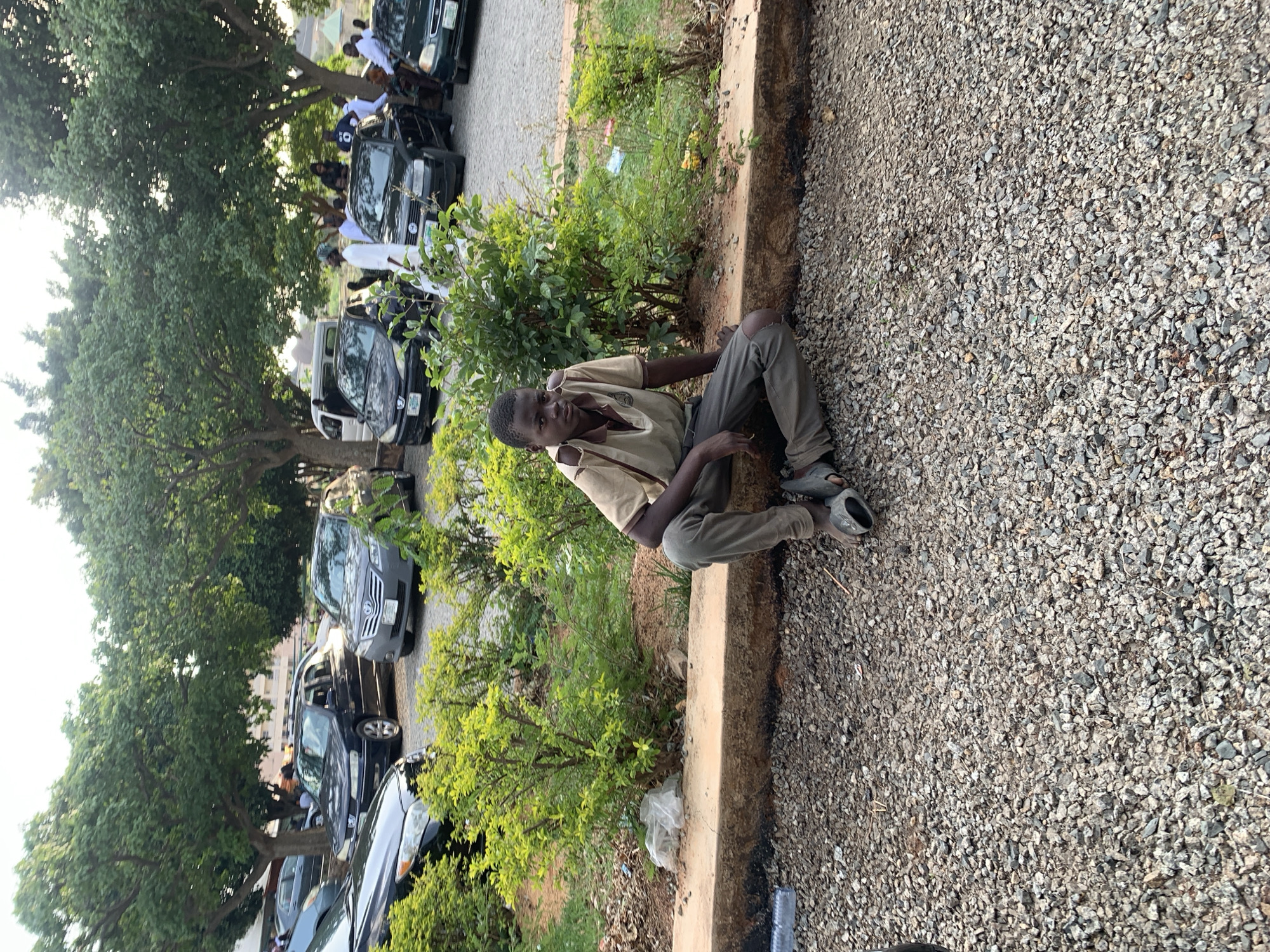 A person is sitting on a concrete curb in Jos, Nigeria. The individual is wearing a tan short-sleeved shirt, gray pants, and open-toed shoes. The person is seated in a crouched position looking directly at the viewer. Adjacent to the curb is an area covered in small pebbles and stones. On the other side of the curb, there is a small patch of green vegetation, including bushes and grass. Behind the vegetation, there are several cars parked along the side of the road, obscured partially by a large tree on the left of the image. There are other people near the vehicles, dressed in white outfits, but they are at a distance. A discarded plastic bottle lies in the pebbles. The lighting in the image suggests it is taken during the day, likely in the late afternoon or early evening.