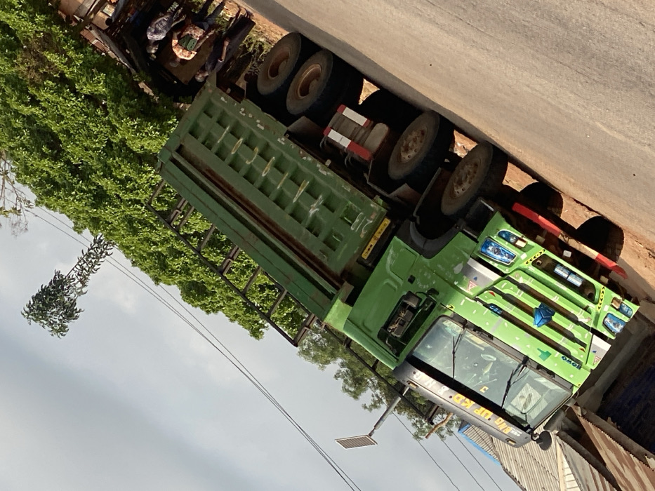 The image captures a roadside scene in Okura, Nigeria, centered on a large, green HOWO brand dump truck. The truck is positioned on an unpaved, light brown dirt or gravel road, facing toward the left side of the frame with its front angled upwards. The truck features a multi-axle configuration, with at least three visible rear axles fitted with dual wheels. Markings on the sun visor read "RNG UP KD RHO," and reflective red and white striped tape is present on the rear chassis.
To the immediate left of the truck's cabin, part of a building with a corrugated metal roof is visible. A solar-powered streetlight on a pole stands nearby. The background behind the truck is dominated by dense green foliage, likely trees or bushes. A taller, slender tree with sparse leaves extends above the main foliage line against a hazy, light grey sky.
On the right side of the image, near the rear of the truck, a group of approximately three to four individuals is visible standing on the dirt shoulder beneath the trees. They appear to be engaged in a stationary interaction or observation. One individual is identifiable by a patterned shirt. The overall lighting suggests daylight conditions.