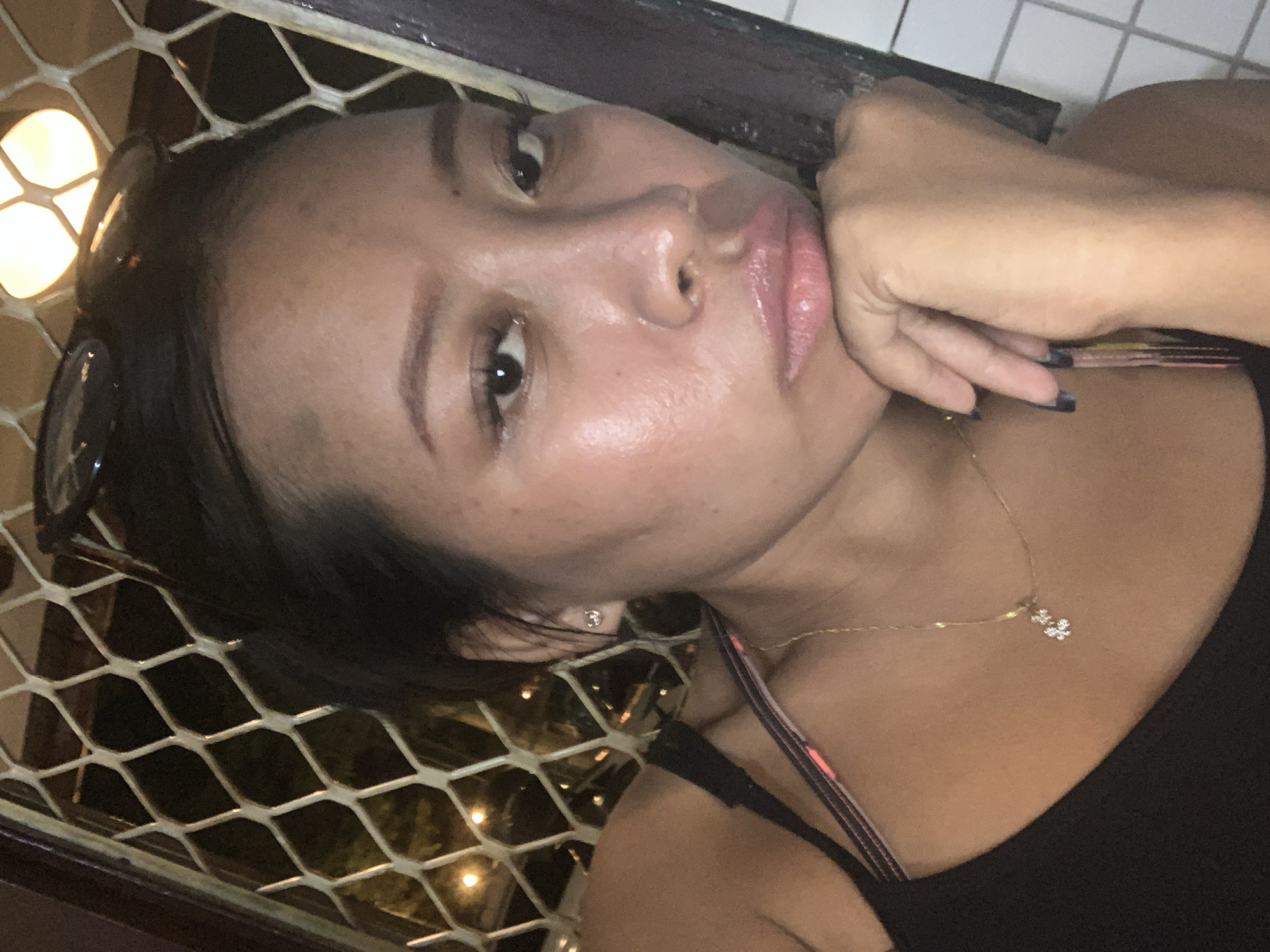 A person is seen in a close-up, angled self-portrait. The person's head is turned slightly to the side with one hand resting near their face. Details like an earring and a necklace are visible. The background features a grid-like screen, partially obscuring a dimly lit area, possibly an outdoor scene. The picture was taken in Tamanbaru, Indonesia.