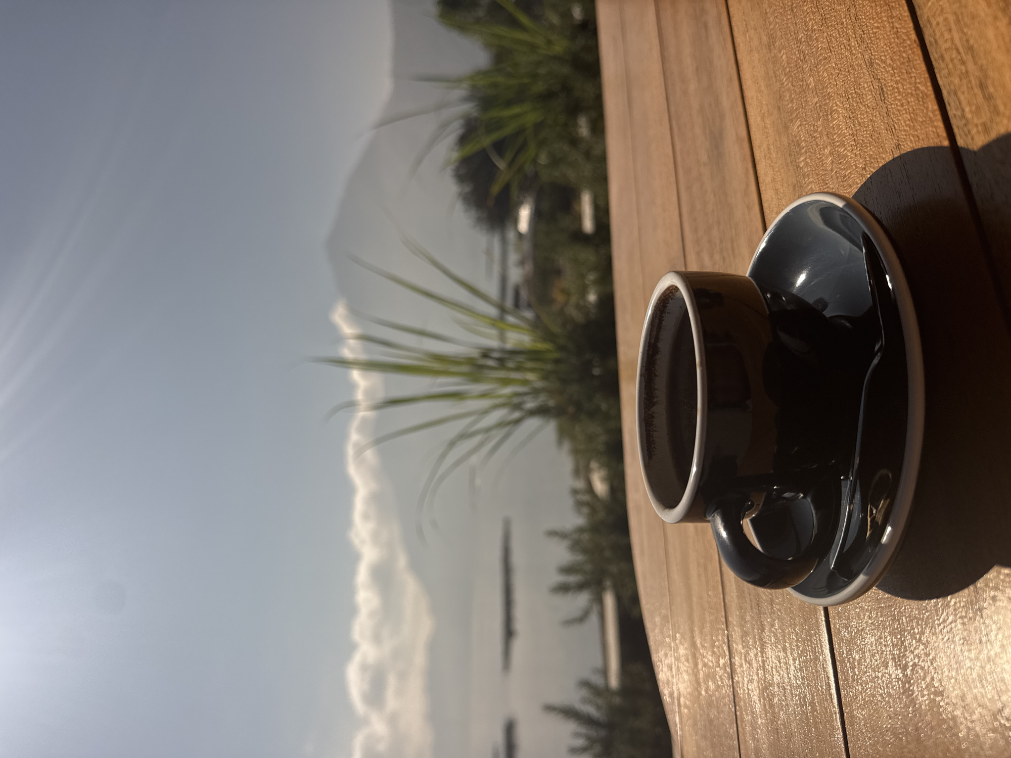 A dark brown ceramic espresso cup and saucer sit on a wooden table. A dark liquid fills the cup. The background is a hazy landscape of a body of water, mountains, and vegetation, under a bright sky with sun flares. The scene is located in Kintamani, Indonesia.
