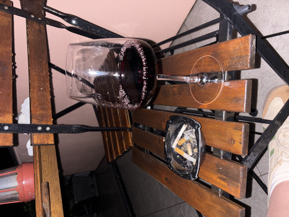 The image depicts a nighttime outdoor scene, viewed from a low angle, focusing on a wet surface with two primary objects. A clear, stemmed wine glass, approximately half-full of dark red wine with visible bubbles around the liquid's surface, stands on a wet, slatted wooden surface. To its left, a black, square ashtray contains four cigarette butts and a quantity of ash and debris. Both the wooden slats and an underlying dark metal frame exhibit numerous water droplets, indicating recent precipitation or heavy condensation.
In the upper left background, another higher, wet wooden surface is partially visible, holding a red, cylindrical object resembling a lantern or small heater, along with a small patch of light-colored, possibly frozen, material. A blurry, light pink wall or building facade forms the main background, with darker, indistinct shapes below the upper wooden surface. In the lower left corner, a partial view of a human leg and foot is visible, wearing light-colored footwear with a textured sole and what appears to be a light-colored sock or garment hem.
The image contains no discernible landmarks or specific visual cues to identify the location as the city of Nesso, Italy.