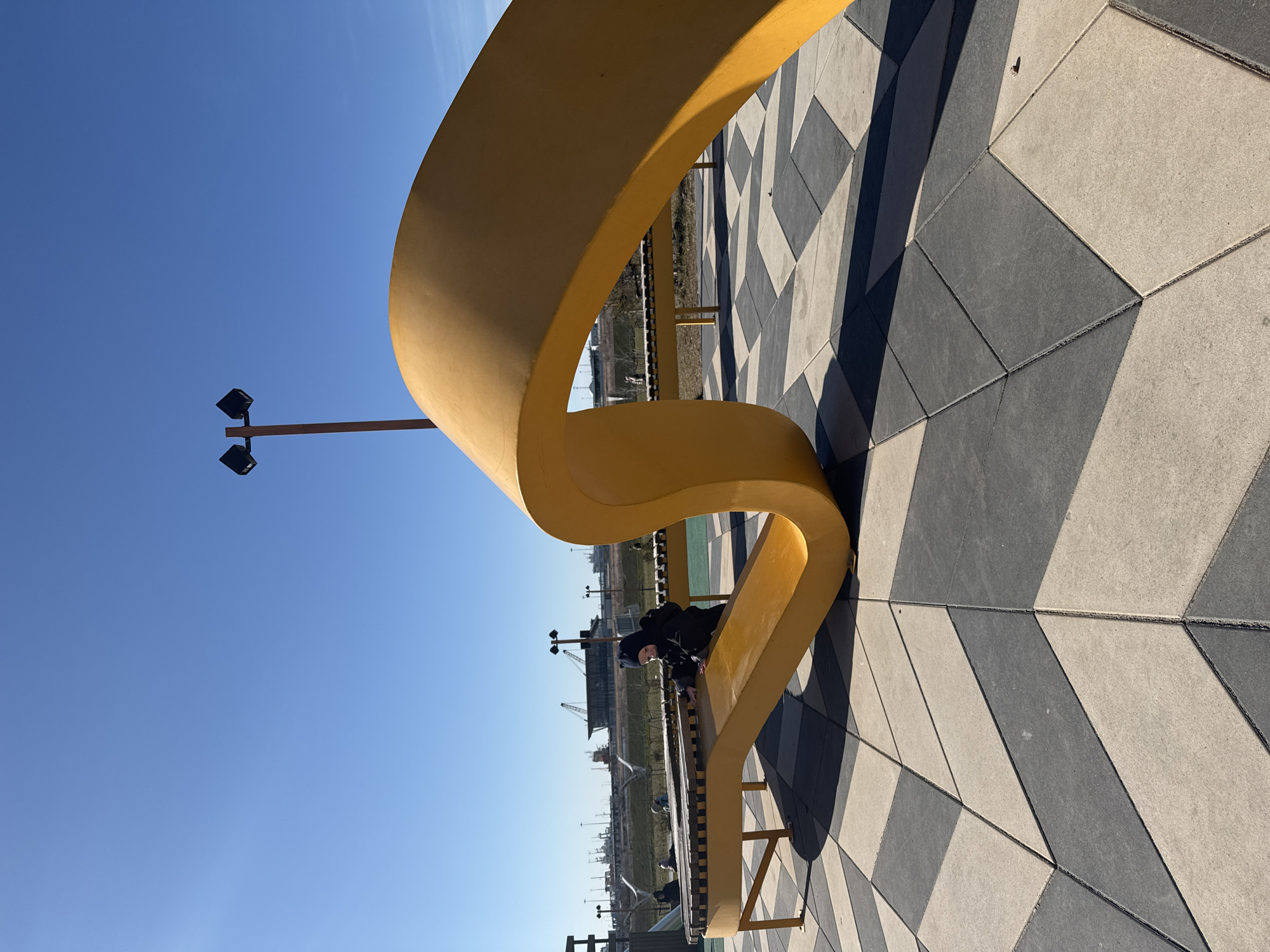 A low-angle, color photograph depicts a yellow, curvilinear bench with a child sitting on it, set in an urban environment under a clear blue sky. The bench, which is the dominant feature, has an S-curve design and is placed on a surface patterned with alternating light and dark gray geometric shapes.
A child is seated on the bench, dressed in a dark, full-body garment with a hood, and is positioned towards the center of the bench's curve.
In the background, the scene shows a cityscape with industrial elements like cranes and infrastructure, suggesting a port or industrial zone. A light pole with two lamps is visible near the bench.
The photograph has a slight tilt. The scene is identifiable as the "Bench of Reconciliation" on Vasilyevsky Island, Saint Petersburg, Russia.