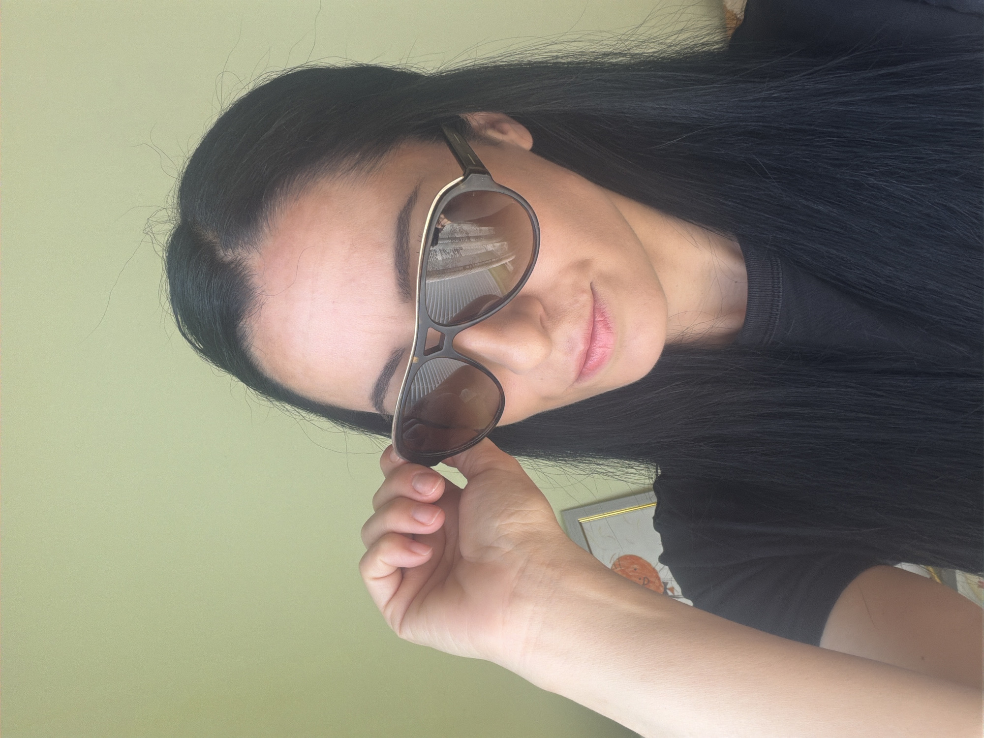 A person with long, black hair is holding sunglasses in their right hand. They are wearing a black shirt. The background is a light green wall. The reflection in the sunglasses shows a building with vertical lines, possibly blinds or a window. Based on the provided information, the location is stated to be in the city of Pleven, Bulgaria, though nothing in the image specifically confirms this.
