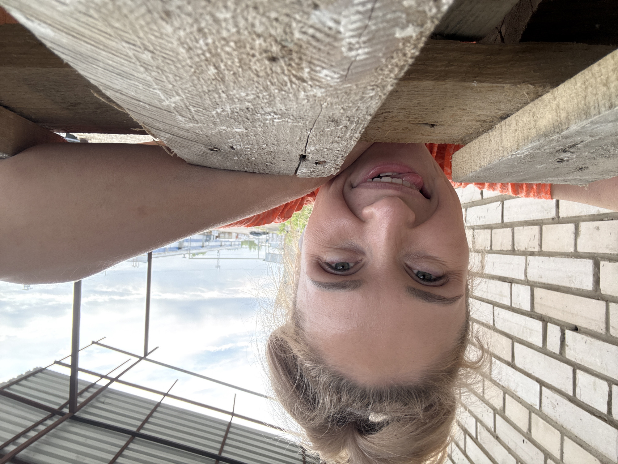 A fair-skinned woman is hanging upside down, positioned between weathered wooden planks and a brick wall. She is smiling with her tongue sticking out. The background shows a glimpse of a cityscape under a cloudy sky and the frame of a metal structure. The location may be Chaikovsky, Russia, as that was specified in the prompt.