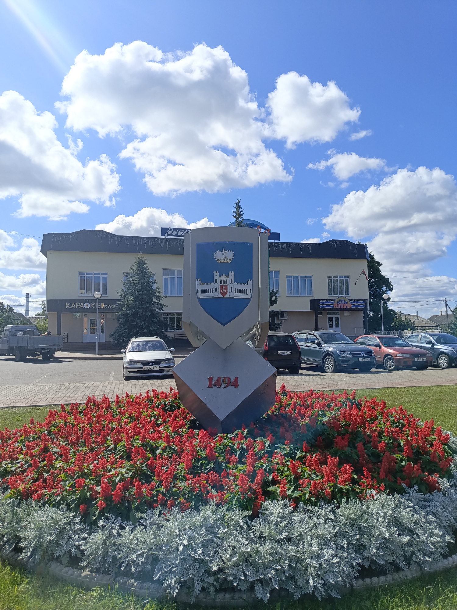 A medium shot captures a sunlit scene in Ivatsevichy, Belarus, dominated by a large, multi-tiered flower arrangement featuring bright red salvia and silvery-white foliage. At the center of the arrangement is a stone monument with a coat of arms depicting a castle and a crown, along with the numerals "1494." Behind the monument and flower bed, a light-colored, two-story building stands, with a sign reading "КАРАОКЕ КАФЕ" visible on the left side and "РЕСТОРАН" on the right. A number of cars are parked in front of the building, including a silver Peugeot with a Belarusian license plate. A small evergreen tree is planted near the entrance of the building. The sky above is a bright blue with fluffy white clouds, and a faint sunbeam can be seen in the upper left corner. The scene is composed and orderly, with no people clearly visible.