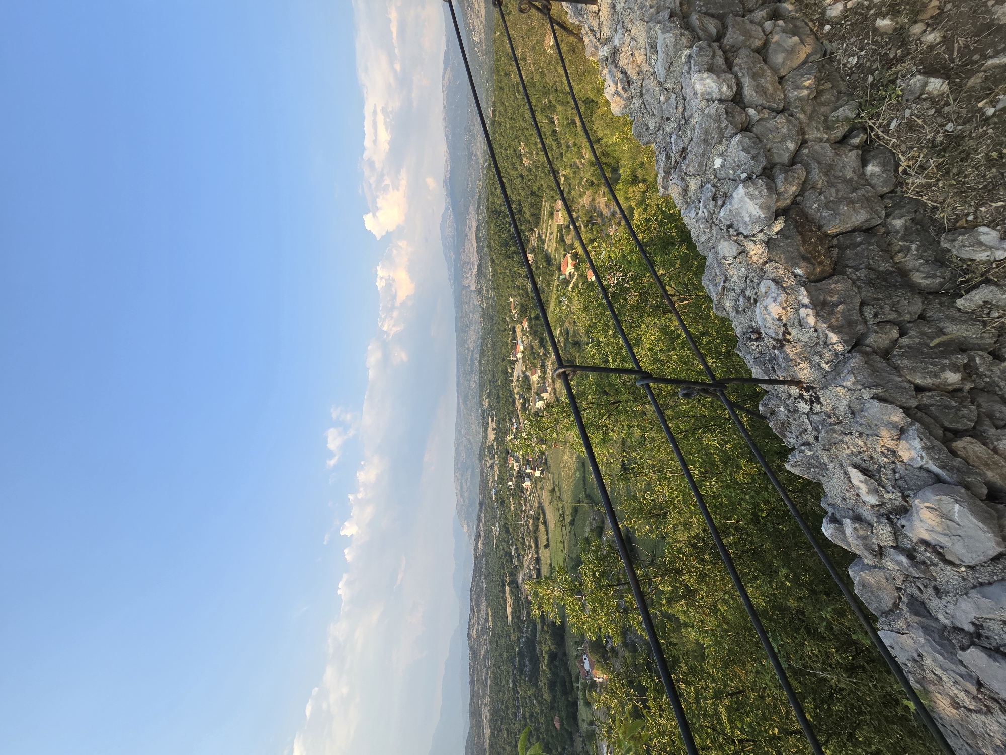 The image depicts a panoramic view of the landscape surrounding Medun, Montenegro, seen from an elevated vantage point. In the foreground, a rough stone wall, likely part of a historical structure, forms a boundary. A black metal railing runs along the top of the wall, offering a protective barrier. Beyond the wall and railing, lush green trees and vegetation dominate the middle ground. In the distance, a rural village with scattered houses featuring red-tiled roofs is nestled within the rolling hills. The landscape extends to a horizon of mountains under a clear blue sky with scattered white clouds. The lighting suggests it is late afternoon or early evening, with soft shadows cast across the scene. There are no people or animals visible in the image.