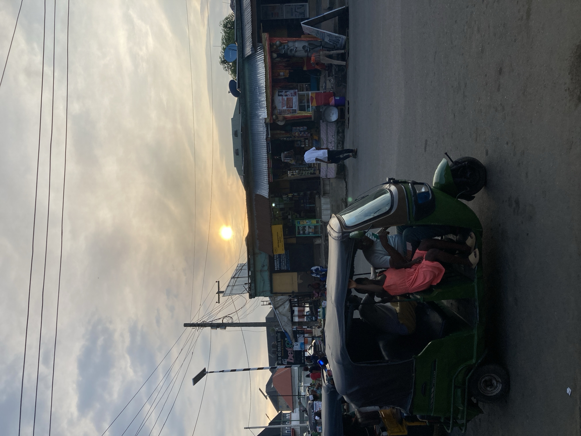 The image depicts a street scene in Aba, Nigeria, at sunset. A green, three-wheeled motorized vehicle, carrying multiple passengers, occupies the foreground. Businesses line the street, with signage visible on the buildings. A person in a white shirt walks near the storefronts. Overhead electrical wires stretch across the sky. The sky is filled with clouds.
