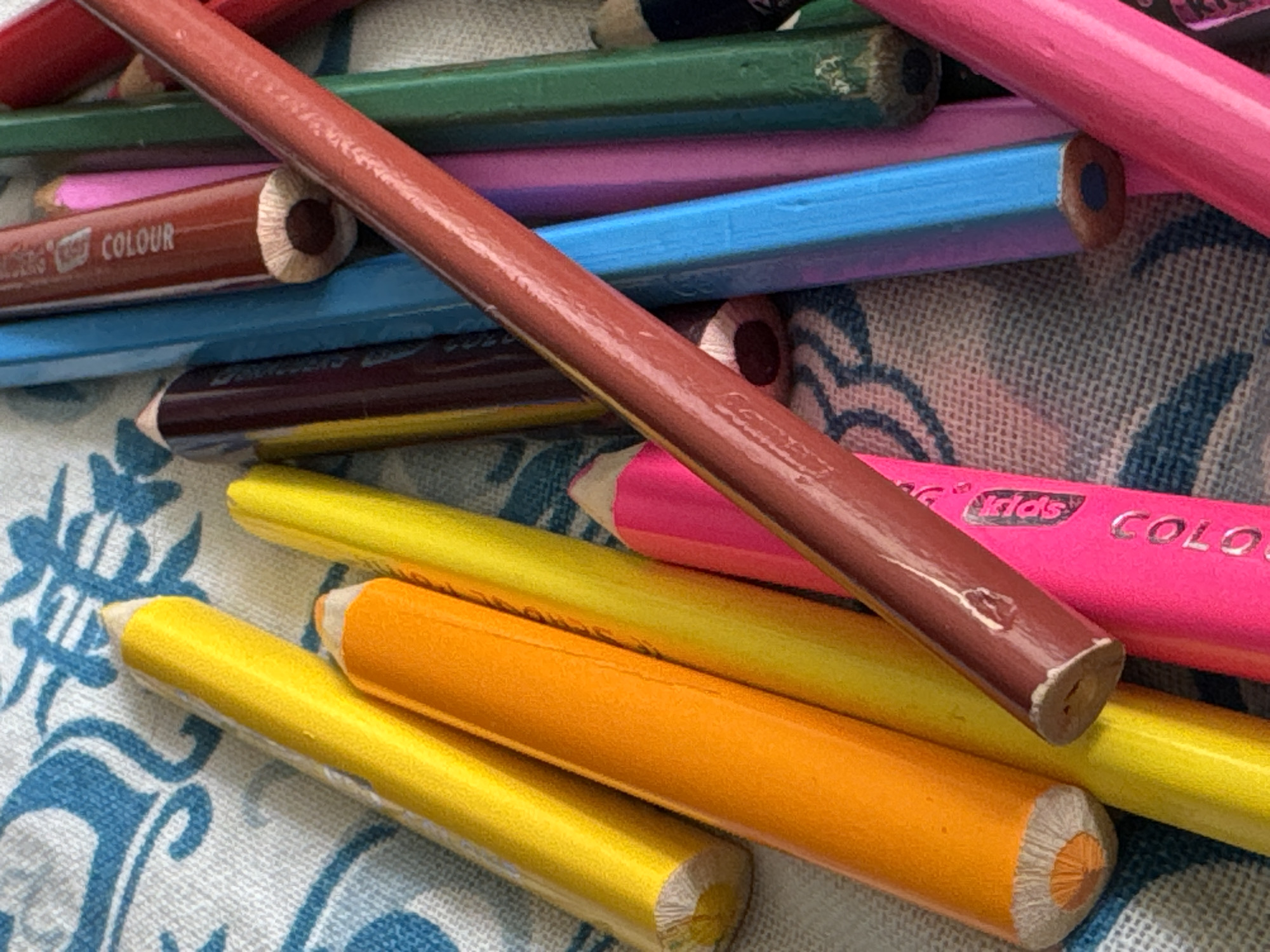 A close-up, overhead shot shows a pile of colored pencils scattered on a white and blue patterned fabric. The pencils are of various colors including yellow, orange, pink, blue, green, brown, and red. Some pencils are sharpened to a point, while others appear to be used. The fabric has a decorative, possibly floral, pattern. The scene is indoors, with no visible windows or external elements. There are no people or animals present. The lighting is even, suggesting it could be artificial. The specific location in Яромінскі сельскі Савет, Belarus cannot be determined from the image.