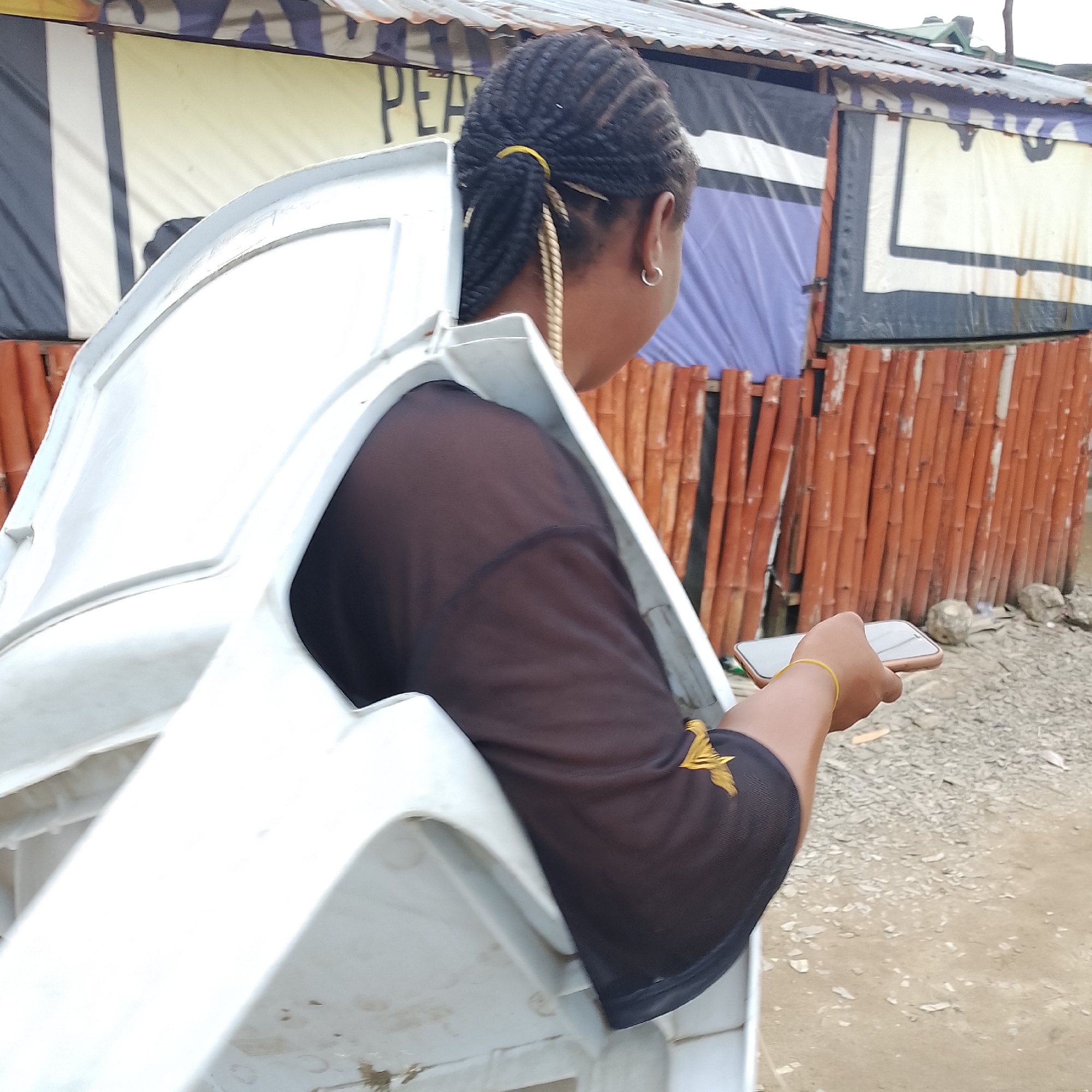 In Ado, Nigeria, a person with braided hair tied with a yellow band carries a white plastic chair, with the chair obscuring much of their body. They are wearing a dark, semi-sheer t-shirt with a gold emblem on the sleeve and a silver hoop earring. The person is holding a smartphone in their right hand, its screen facing upwards. Behind them, a rustic structure with a corrugated metal roof is visible, adorned with colorful tarpaulins and bamboo-like poles. The ground appears to be dirt and gravel.