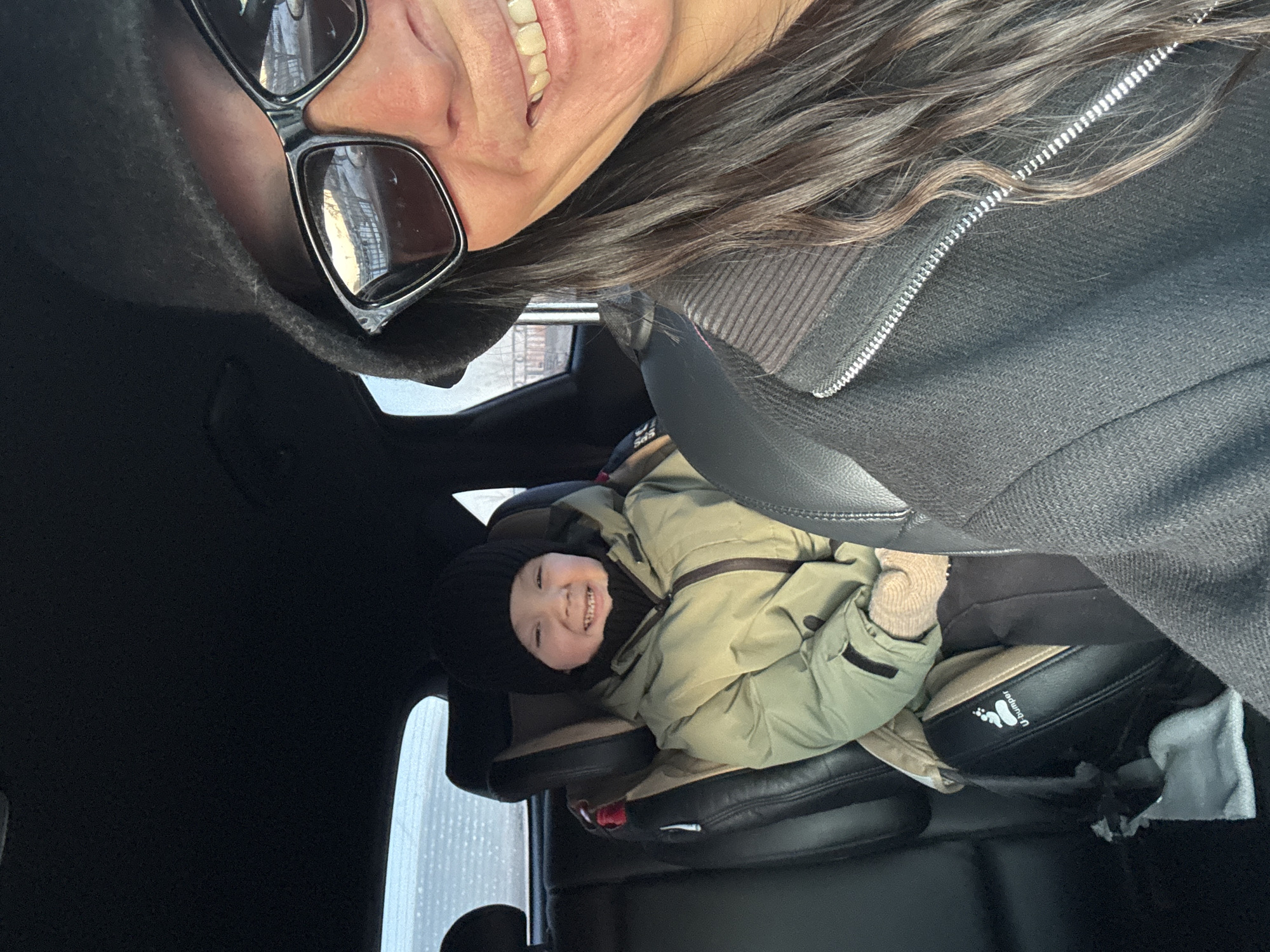 The image depicts a selfie taken inside a car. An adult woman is visible in the upper portion of the image, wearing sunglasses and a dark hooded jacket. Below her, a child is seated in a car seat, wearing a dark hat and a green jacket. The child is smiling. The interior of the car is visible, including the car seat and a window with what appears to be a rainy or snowy view outside. Based on the rainy window view, the image was taken in Chaikovsky, Russia.
