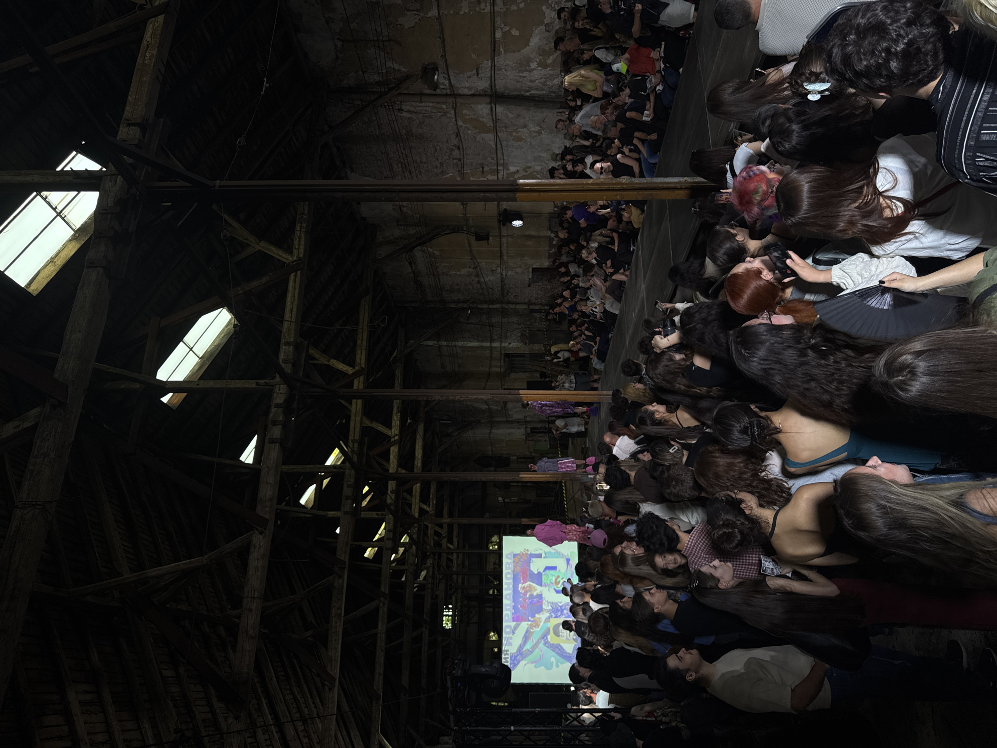 An indoor fashion show is taking place in a large, dilapidated industrial building. The wooden roof structure is exposed and weathered, with skylights allowing natural light to filter in. A runway is set up in the center of the space, illuminated by a large projector screen displaying colorful abstract graphics. Models are walking down the runway, dressed in vibrant, avant-garde outfits. A crowd of spectators, dressed in casual attire, is seated on tiered benches on either side of the runway, with some people standing in the background. Many audience members are holding up their phones, seemingly recording or taking pictures of the event. The overall atmosphere appears to be energetic and fashionable, with a contrast between the raw industrial setting and the contemporary fashion on display.