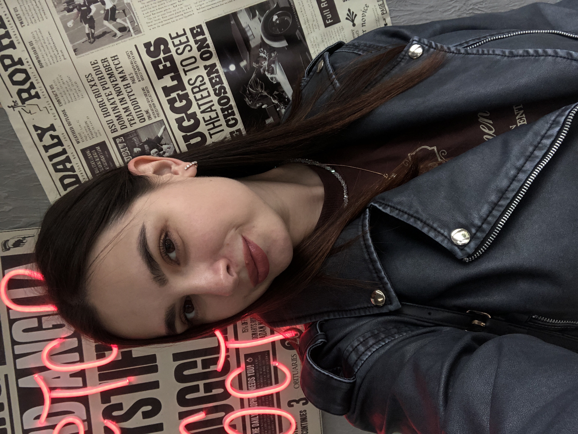 A young woman with long, dark hair and a black leather jacket is posing for a selfie. She is wearing a dark maroon shirt and a necklace. Behind her, there is a wall decorated with what appears to be newspaper clippings and neon signs. The newspaper clippings have headlines related to "The Daily Prophet" and "The Chosen One." One of the neon signs appears to be in Russian and says "Я люблю Ижевск" which translates to "I love Izhevsk." This suggests the location is in Izhevsk, Russia. The lighting is dim, with the neon signs providing a pinkish glow. The woman is looking directly at the camera with a slight smile. The image captures a casual and trendy aesthetic, possibly taken in a cafe or a themed establishment.