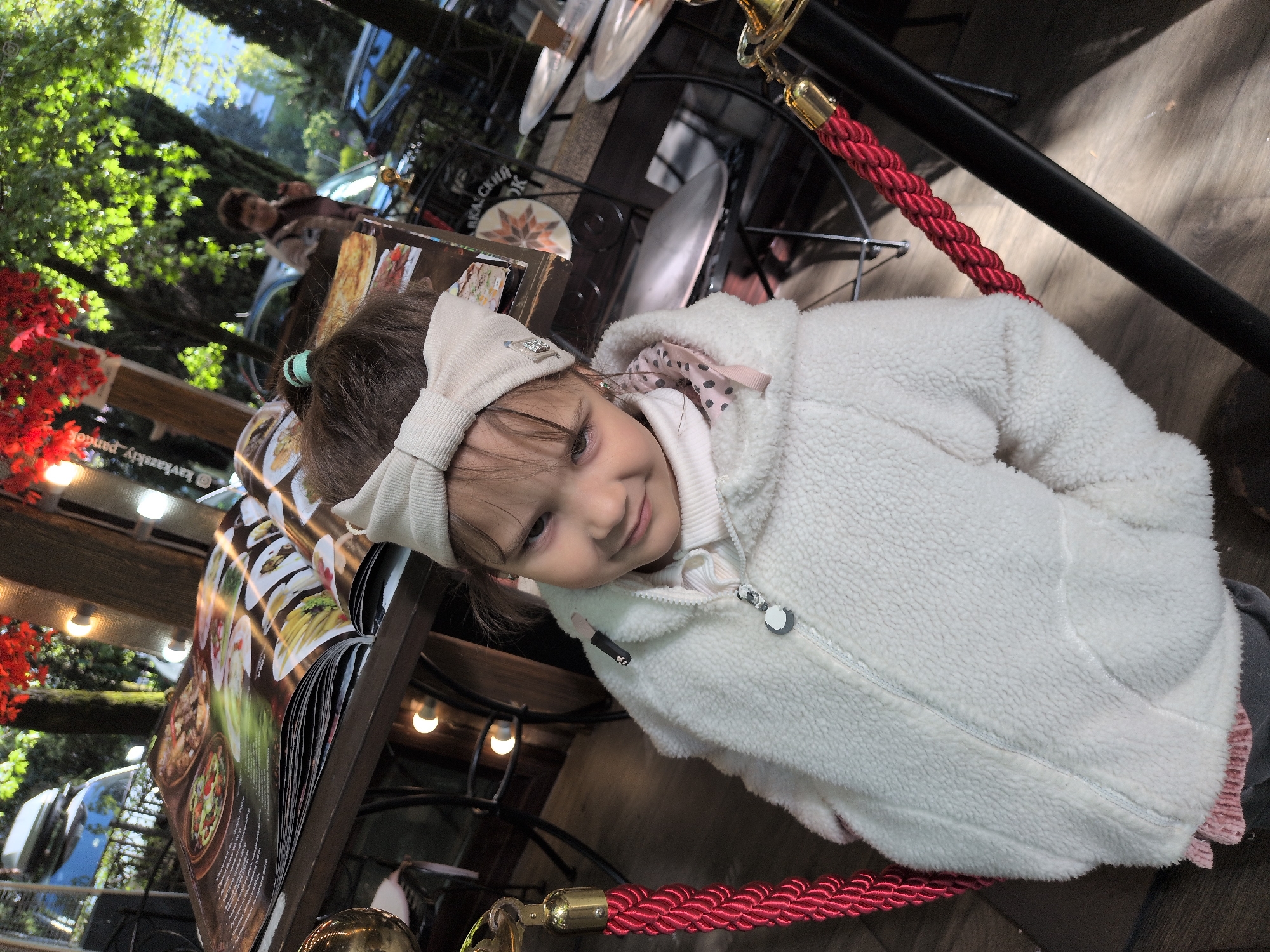The photograph shows a young girl in a white, fleecy jacket and a beige bow headband, seated at an outdoor cafe. She is positioned next to a menu stand that displays images of food. The cafe appears to be located outside, near trees and possibly in a park-like setting. The presence of red rope barriers suggests an organized outdoor space, possibly a cordoned-off seating area. In the background, there is a glimpse of street activity with cars and other pedestrians. The location is in the city of Sochi, Russia.