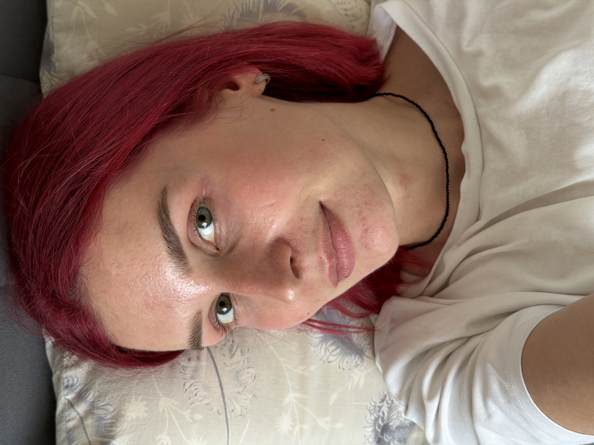 A young woman with striking red hair is depicted in a close-up, high-angle shot, lying down indoors, likely in Chaikovsky, Russia. She is positioned on her side, resting her head on a light-colored pillow with a faint floral or leaf pattern. Her head is turned towards the viewer, and her green or hazel eyes are open, looking directly at the camera with a neutral to slightly serene expression.
She has a fair complexion, and her face shows natural skin texture, including some minor redness and small blemishes. Her eyebrows are dark and well-defined. Her lips are a natural pink hue, appearing soft. A small, sparkly earring is visible in her left ear. She is wearing a simple, loose-fitting white t-shirt, and a thin, black beaded necklace is visible around her neck, resting against her skin. The background behind her head appears to be a dark gray surface, possibly a sofa or bed frame. The lighting suggests daytime, with soft, natural light illuminating her face. There is no visible text in the image.