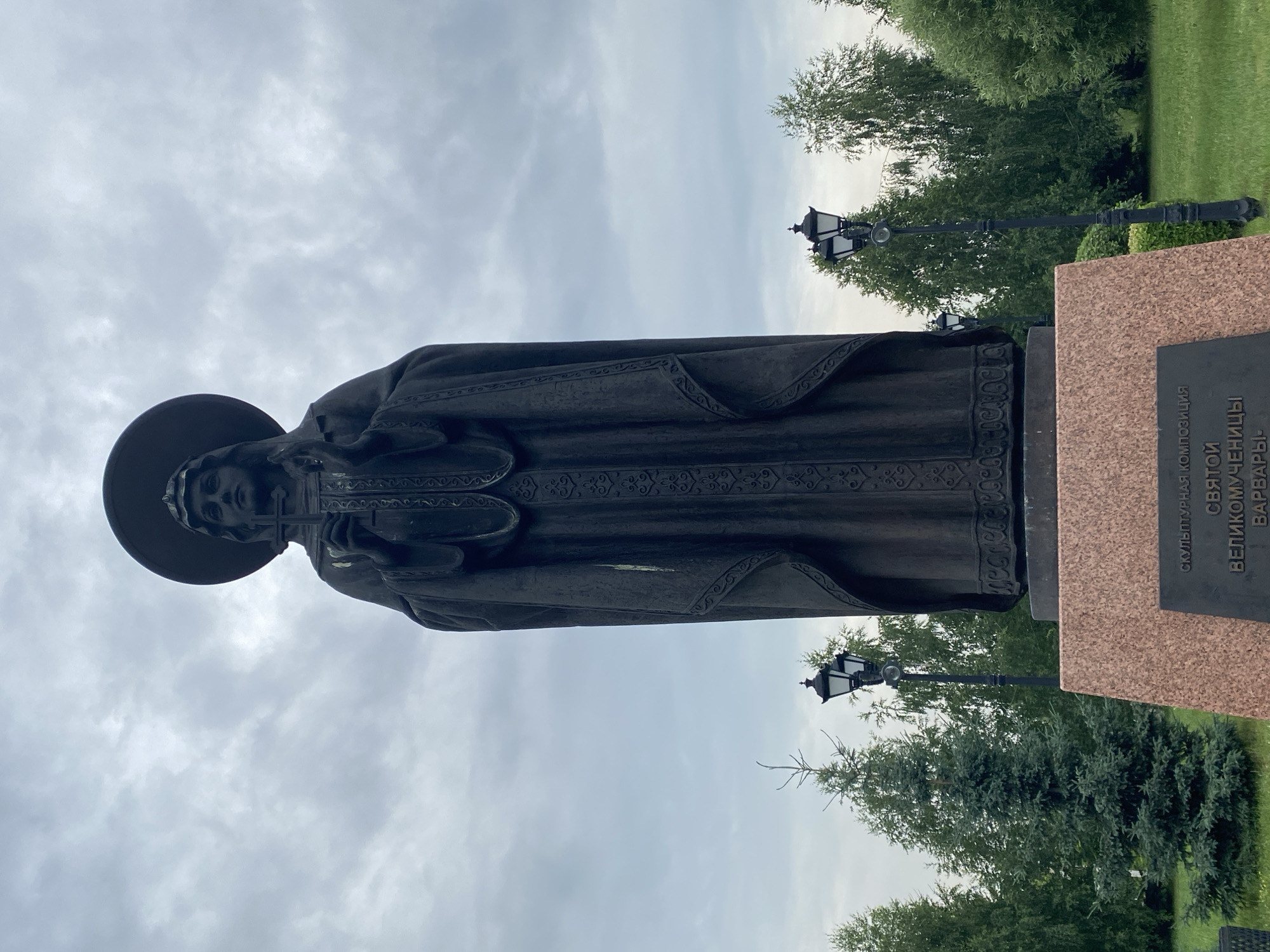 The image shows a dark bronze statue of a woman in a religious robe, holding a cross. She has a halo around her head. The statue is on a pinkish-brown granite pedestal, which has an inscription in Russian. The inscription reads "СКУЛЬПТУРНАЯ КОМПОЗИЦИЯ СВЯТОЙ ВЕЛИКОМУЧЕНИЦЫ ВАРВАРЫ" which translates to "SCULPTURAL COMPOSITION OF SAINT THE GREAT MARTYR BARBARA". The statue is located in a park-like setting with green grass, trees, and two ornate black lampposts. The sky is overcast. This statue is located in Novokuznetsk, Russia, in the Park of the Victory.