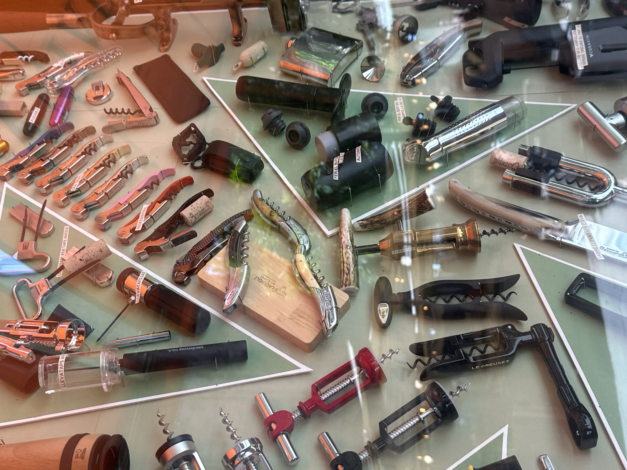 This image is a close-up, eye-level shot of a display window exhibiting a variety of wine openers and related accessories, possibly in Barcelona, Spain, given the visible text. The collection is arranged on a pale green surface with white triangular geometric shapes. The lighting creates reflections on the glass, obscuring some details.
The central focus is a diverse array of corkscrews, ranging from classic waiter's friends and T-handle openers to more modern, automated devices. Several prominent items include a red T-handle corkscrew, a black multi-tool corkscrew labeled "LE CREUSET," and a metallic corkscrew with a clear cylinder, labeled "LLEVATAPS AI." There are also several Laguiole-style corkscrews, identifiable by their distinctive handles and branding.
In addition to corkscrews, the display features wine stoppers, bottle openers, and what appear to be wine preservation systems, such as the "CORAVIN" device with a black body and metallic accents. Some items are presented with wine corks, some still in place. The background is a mix of reflective surfaces and other, less distinct objects, suggesting the display is within a shop or establishment. The overall impression is one of a curated collection for sale, emphasizing functionality and perhaps artisanal quality.