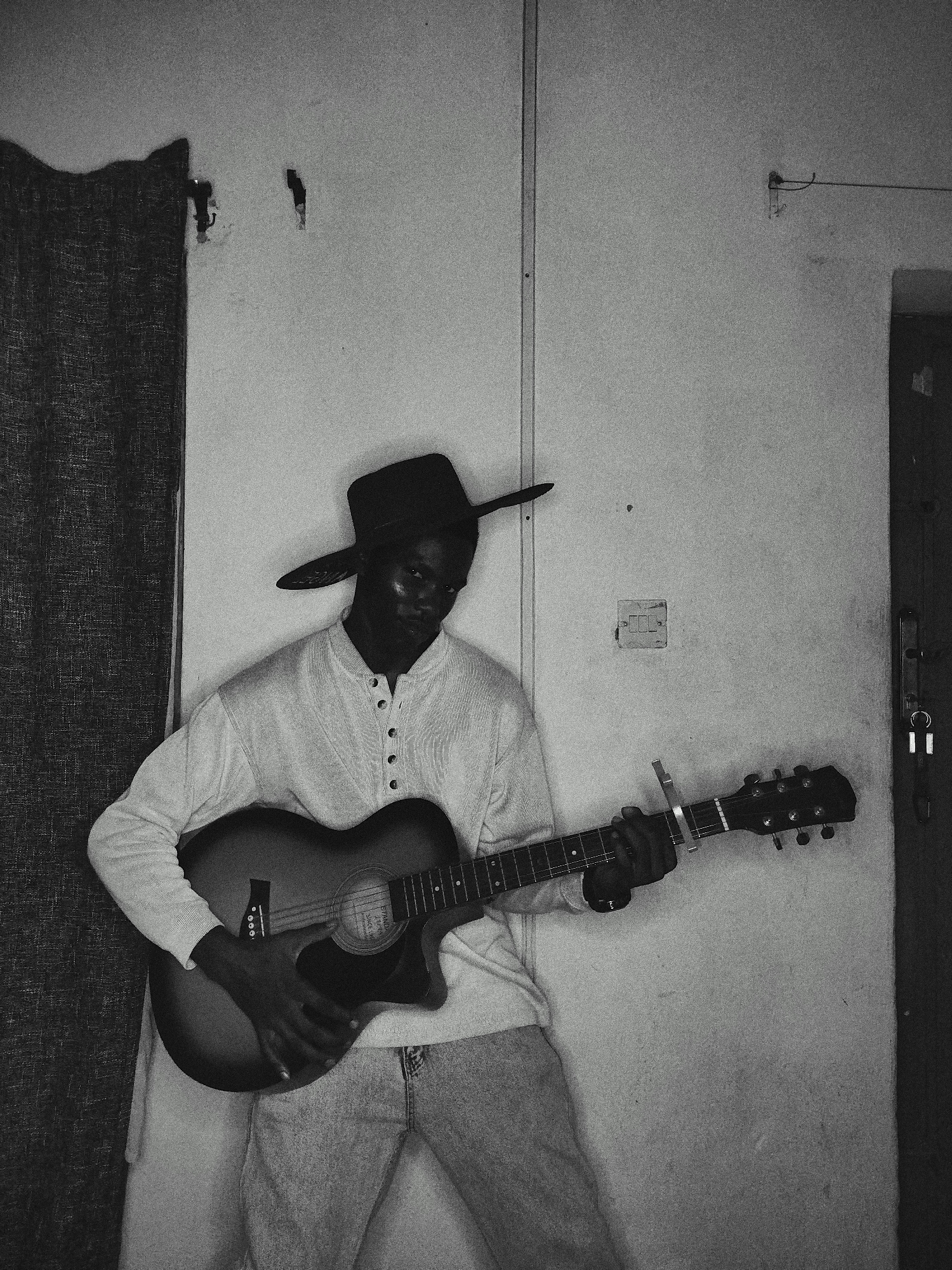 A black and white photograph captures a man playing an acoustic guitar indoors. The man wears a long-sleeved shirt with buttons and light-colored jeans. A wide-brimmed hat obscures the top of his head. His right hand strums the guitar strings, while his left hand holds down chords on the fretboard with a capo attached. He appears to be leaning or positioned in front of a wall.
The backdrop features a wall with a light switch, a partially visible door on the right side, and what seems to be a curtain or draped fabric on the left. Additionally, there are a couple of hooks or objects protruding from the wall above the curtain. The scene is dimly lit, creating shadows and obscuring some details.
There is no visible indication of the location being in a specific city.