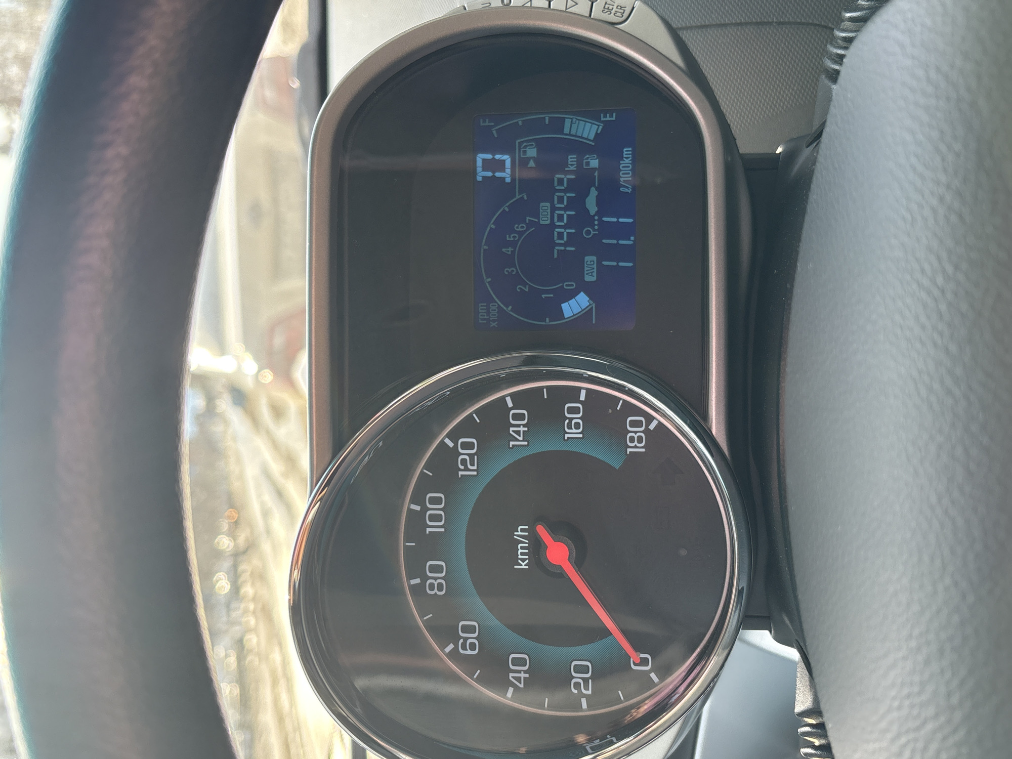 The image captures a close-up view of a car's dashboard from the driver's perspective. The main subjects are the instrument cluster and a portion of the steering wheel.
In the center of the frame is the instrument cluster, which consists of a large analog speedometer below a rectangular digital display. The speedometer measures speed in kilometers per hour (km/h), with markings from 0 to 180. The red needle rests at 0 km/h, indicating the vehicle is stationary.
Above the speedometer, the digital display shows various vehicle information. A digital RPM gauge is visible on the left, marked "rpm x1000", with values from 0 to 7. The fuel gauge is on the top right, indicating the fuel level is approximately half full, between 'E' (Empty) and 'F' (Full). The most prominent reading is the odometer, labeled "ODO", which shows "79999 km", signifying nearly 80,000 kilometers traveled. Below the odometer, the average fuel consumption is displayed as "AVG 11.1 l/100km". A gear indicator shows "D", suggesting the car is in Drive mode.
Parts of the steering wheel are visible on the left and right sides of the image, framing the dashboard. The background, seen through the windshield, is blurred but appears bright, suggesting it is daytime. On the top right of the dashboard, beyond the digital display, some control buttons are visible, including "SET" and "CLR". The setting is the interior of a car, likely in Ust-Ilimsk, Russia, given the provided context and the use of metric units for distance and fuel consumption.