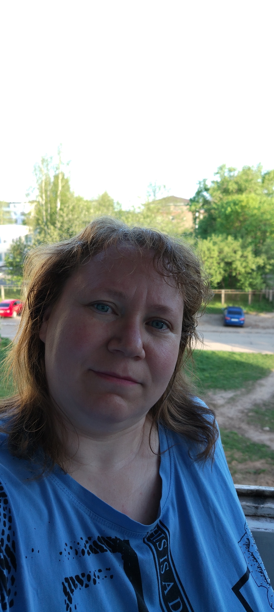 The image shows a middle-aged woman in a blue T-shirt with black lettering on it. The background indicates an outdoor setting, likely in a residential area. There are trees, a building, and a dirt area with two cars parked. The general location appears to be in a city, possibly Chaikovsky, Russia, given the prompt. The image is a close-up shot of the woman, with the background slightly out of focus.