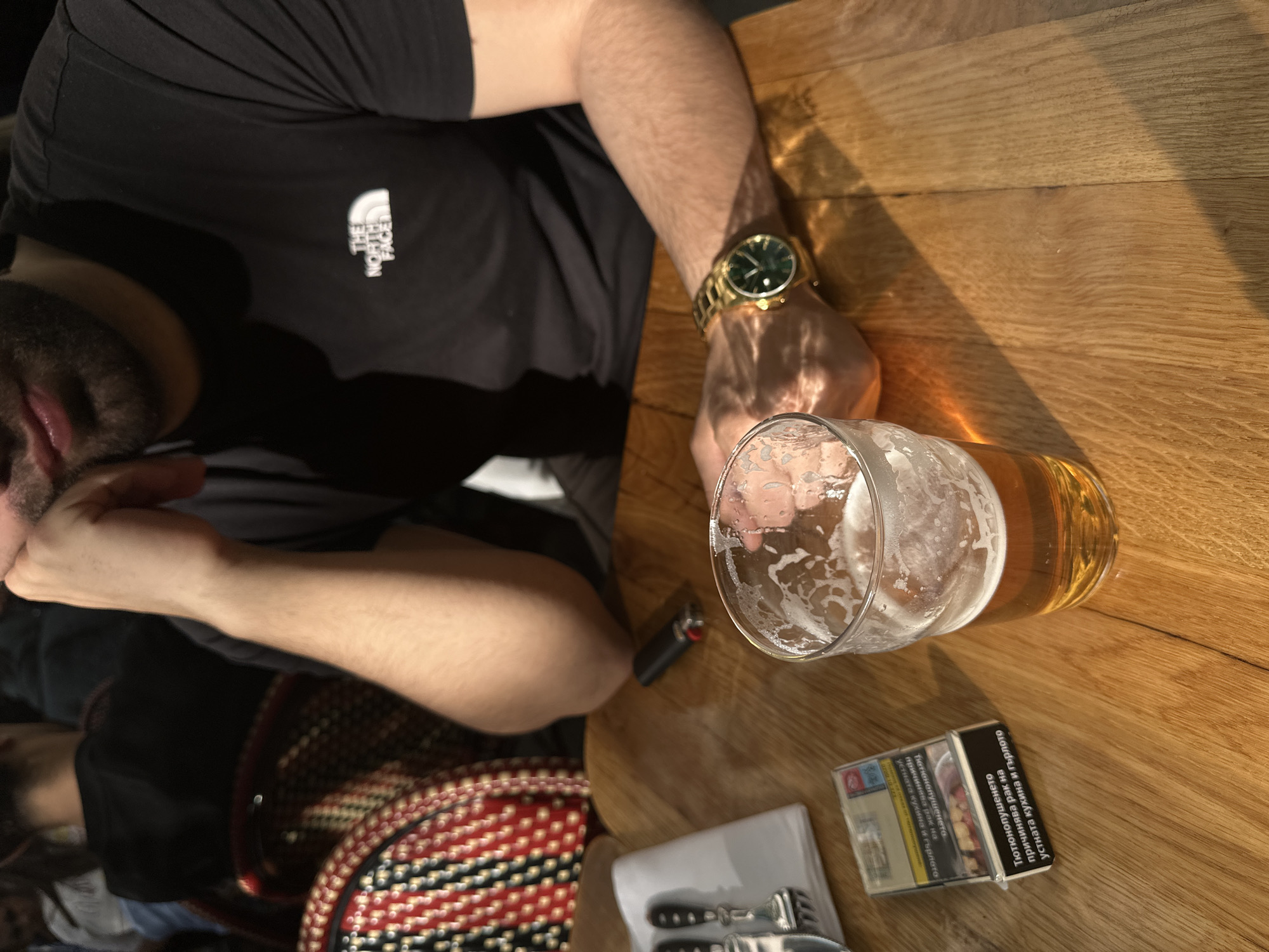 A photograph, taken from a high-angle perspective, shows a man seated at a wooden table in Belgrade, Serbia. A partially consumed glass of beer sits on the table next to a pack of cigarettes, a lighter, a paper napkin, and some cutlery. The man wears a black t-shirt with "The North Face" logo and a gold watch. The table surface is oak wood, illuminated by natural light.