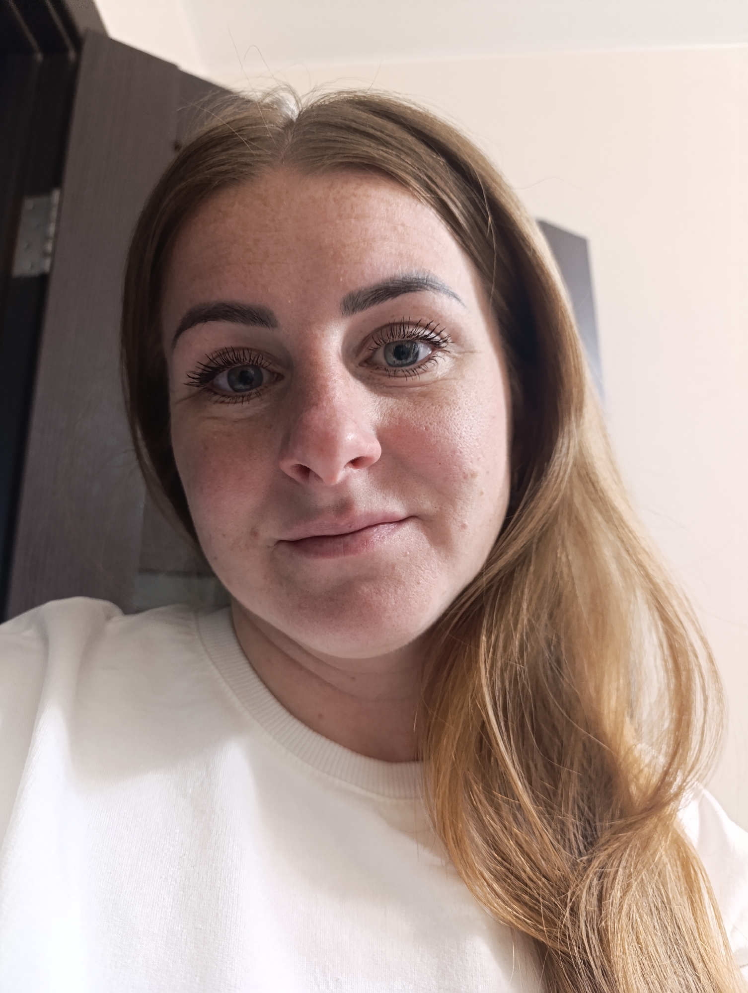 A close-up selfie shows a Caucasian woman with fair skin, freckles, and reddish-blonde hair. She has blue eyes and appears to be wearing mascara. She is wearing a white sweatshirt. The background includes a dark brown door frame on the left and a light beige wall on the right. The location and city are unknown.
