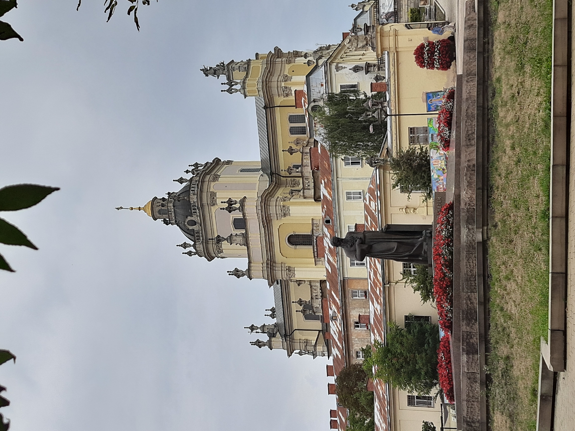 The image shows the St. George's Cathedral in Lviv, Ukraine, a prominent landmark of Ukrainian Baroque architecture. The cathedral is a large, yellow building with a prominent dome and ornate decorations. In front of the cathedral, there is a statue of a religious figure standing on a pedestal. The statue is dark in color and appears to be made of bronze or stone.
The foreground of the image features a well-maintained lawn and a stone embankment. Red flowers are planted in rows along the embankment, adding a splash of color to the scene. Several people can be seen in the background, some walking and others standing. The sky is overcast, suggesting a cloudy day. The overall atmosphere is serene and historic, capturing the grandeur of the cathedral and its surroundings.