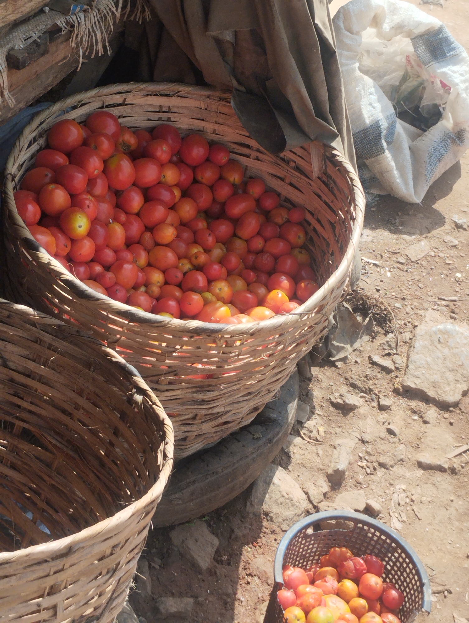 This image shows a street market in Dutsen Alhaji, Nigeria. The foreground features three baskets of tomatoes. The largest basket is overflowing with red and orange tomatoes. A second, similar basket is partially visible on the left. A smaller, blue plastic basket containing more tomatoes sits in the lower right corner of the frame. The baskets are sitting on the ground on what appears to be dirt or cracked pavement. Behind the largest basket, there is a partially visible bag, and the surrounding area appears to be a bustling market scene. The lighting is bright, suggesting it's daytime. There are no people visible in the frame.