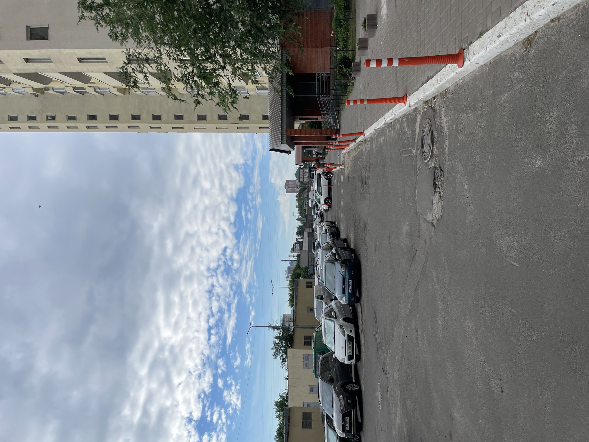 The image depicts a street scene in Kyiv, Ukraine, with a tall, light-colored apartment building on the right. The building has many windows and balconies. In front of the building, there is a covered entrance with red brick walls. Orange and white traffic cones line the sidewalk, which is paved with gray tiles. Adjacent to the sidewalk is a parking lot filled with various cars, including a dark gray SUV, a white hatchback, and a blue sedan. Beyond the parking lot, a row of low-rise buildings with light-colored facades and dark windows can be seen. The sky is cloudy with patches of blue. The ground in the foreground is asphalt with some cracks and a manhole cover. A tree with green foliage is visible near the apartment building. There are no people clearly visible in the image, although a figure might be present near the entrance of the building on the right. The scene appears to be an ordinary urban residential area.