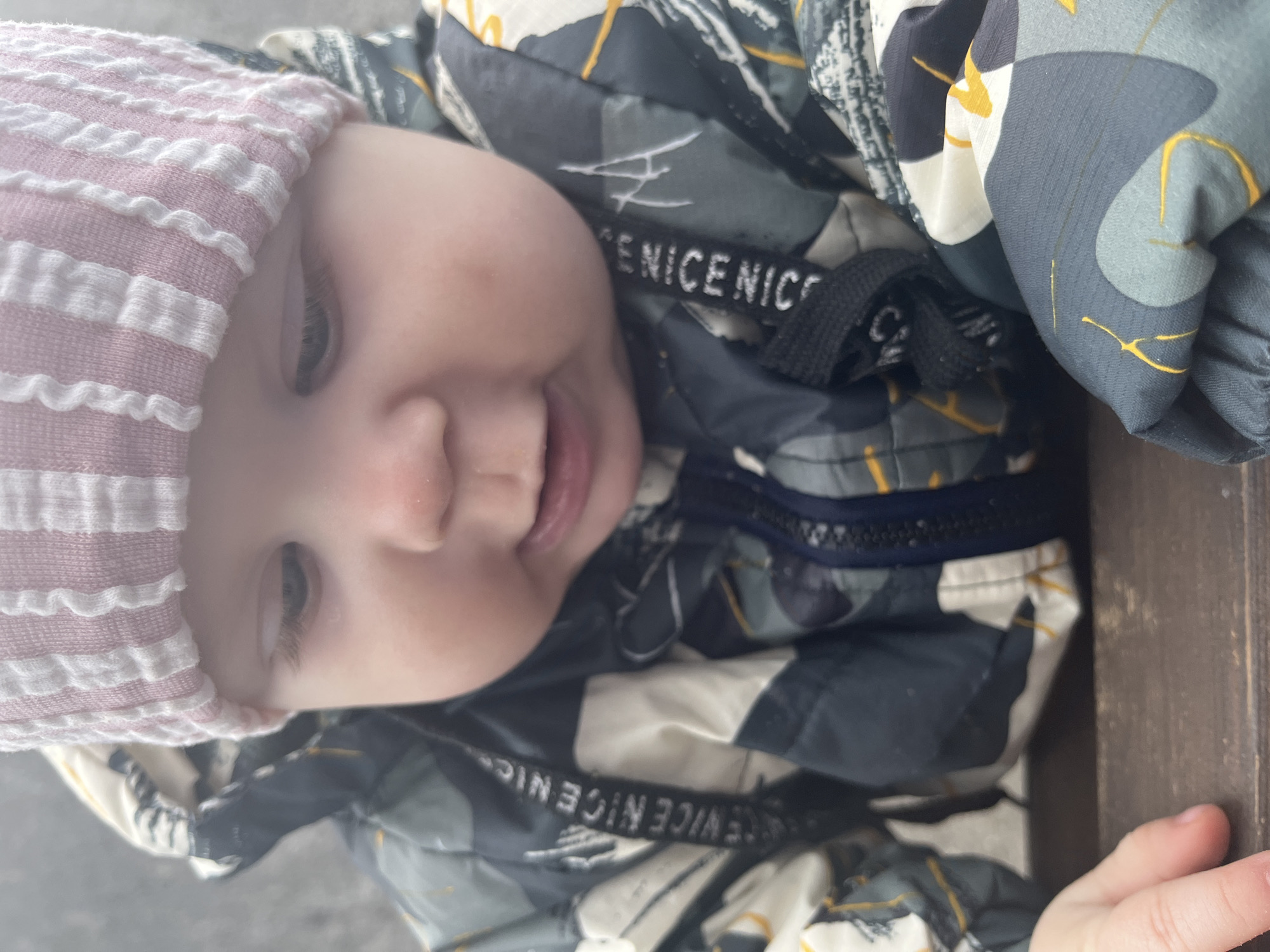 A close-up photograph displays an infant, estimated to be between six and eighteen months old, positioned at eye level. The infant wears a light pink and white striped knitted beanie and a padded winter jacket featuring an abstract pattern in shades of dark green, grey, white, and yellow. A black drawstring with "NICE NICE NICE" text is visible on the jacket's left side. The infant's face shows round cheeks, slightly parted lips, and eyes directed downwards and to the left of the frame. Faint red marks are present on the infant's left cheek. The infant's left hand rests on a dark brown, grainy wooden surface that occupies the lower portion of the image. The background is uniformly light grey and out of focus, providing no discernible environmental details. The specific location within the city of Sochi, Russia, cannot be determined from the visual information provided.