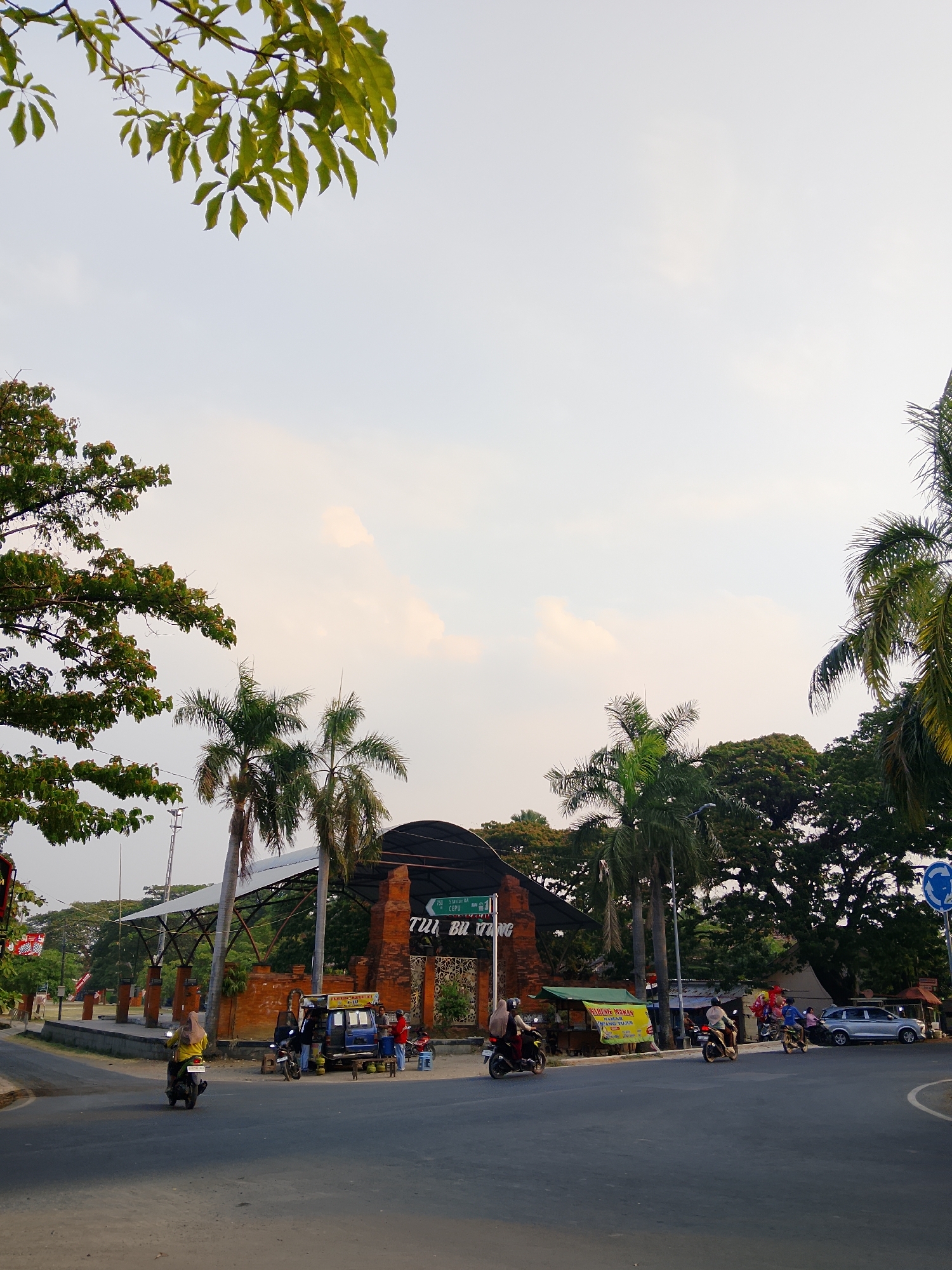 The image depicts an outdoor scene in Balun, Indonesia, likely during late afternoon given the lighting. In the foreground, a paved road curves through the scene. Several motorcycles are visible, with one carrying a person wearing a yellow hijab in the left lane, another with two passengers in the center lane, and one with a single rider in the right lane. Further down the road, a blue vehicle, possibly a van or truck, is parked, with several people standing around it. Adjacent to this vehicle is a food stall with a yellow awning.
Dominating the mid-ground is a structure with a brick facade and a dark, curved roof supported by a metal frame. Above the brick archway, text reads "TUBU BU TRUNG" with smaller text above and below. Tall palm trees surround this structure, and lush green trees form a dense canopy in the background. A white street light is visible behind one of the palm trees.
In the background, more trees and a light pole are visible. A blue circular sign with arrows indicating a roundabout is positioned on the right side of the road. The sky is a pale, hazy blue with a few soft clouds, suggesting fair weather. The overall impression is a typical street scene in a town or city, with local commerce and transportation taking place under a warm sky.
