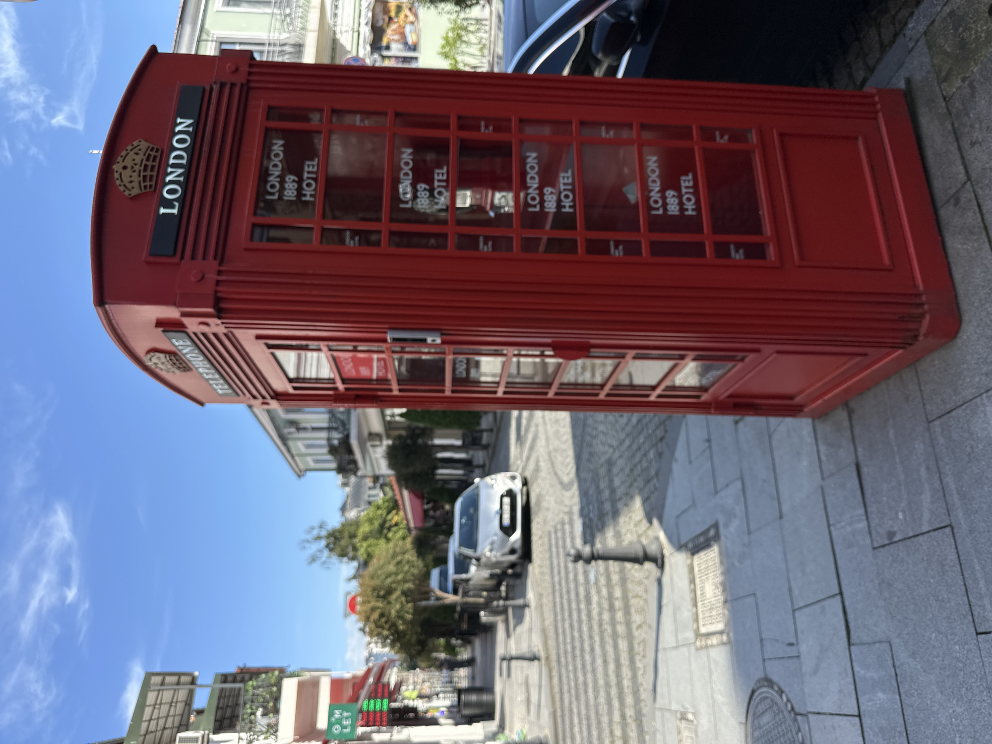 Here's a detailed description of the image:
The image showcases a red telephone booth, reminiscent of those found in London, but located in Batumi, Georgia. The booth is positioned on a paved pedestrian walkway, constructed from grey stone slabs. It's primarily red with "LONDON" vertically printed near the top, as well as the words "LONDON 1889 HOTEL" within the glass windows.
In the background, a street scene is visible. There are buildings, including some with balconies and awnings, lining the road. Cars are parked along the street, and trees provide greenery to the scene. The sky is blue, suggesting a clear day.